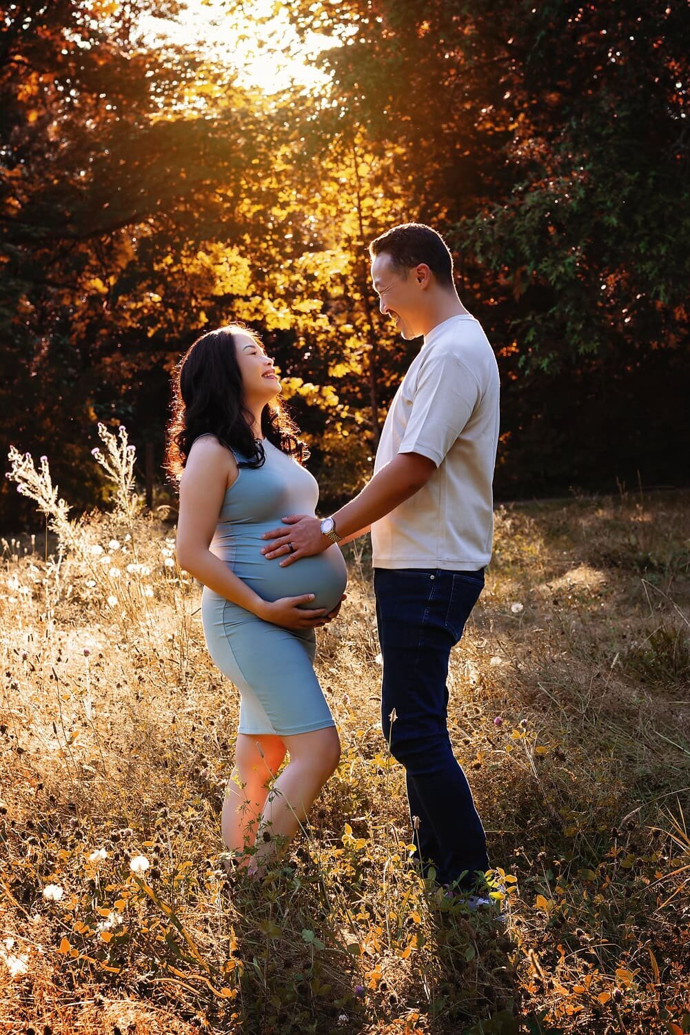An expecting couple in a field at Deer Lake Park in Burnaby, with the mother in a blue dress during an outdoor maternity session at sunset.