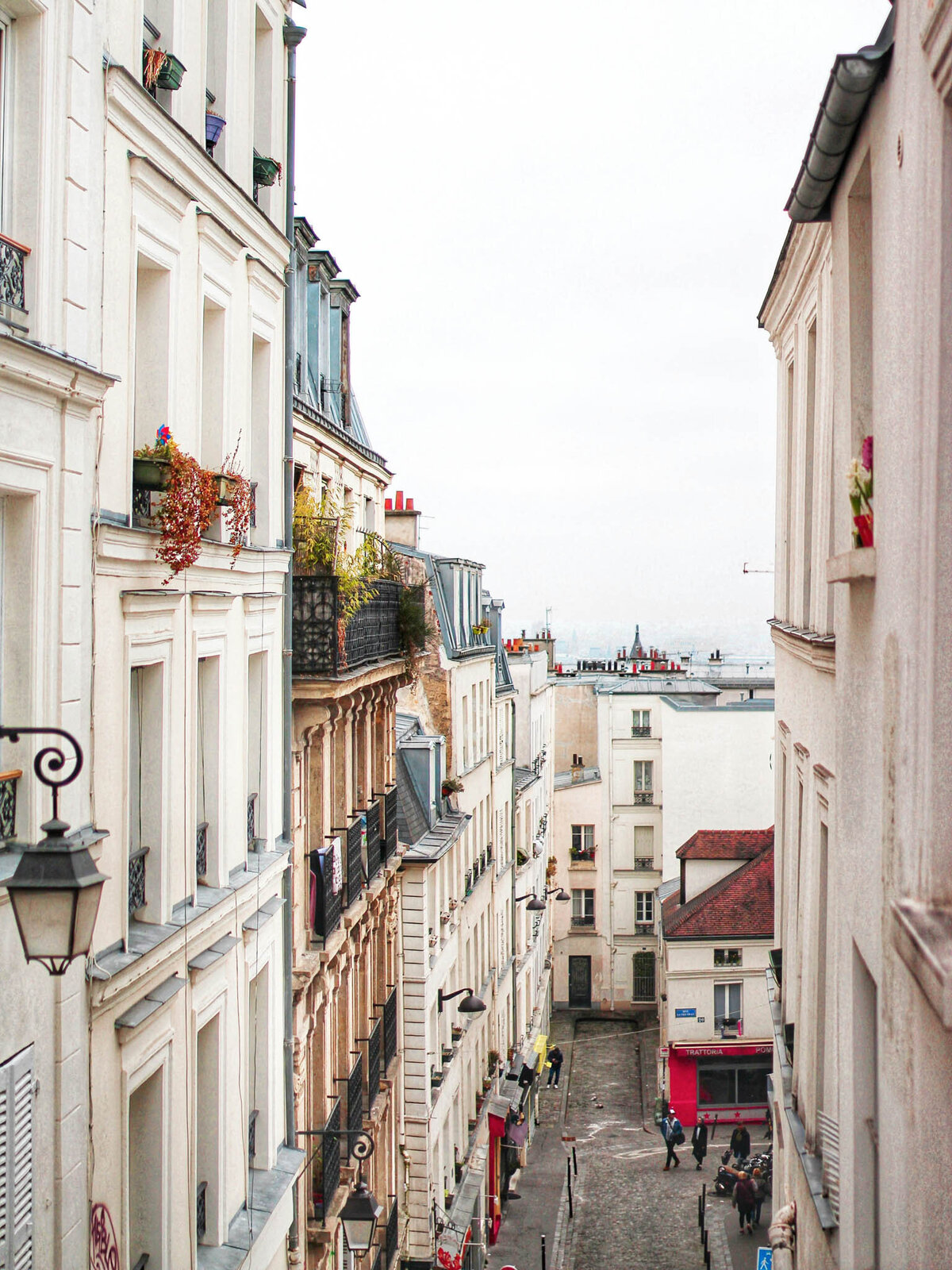 A narrow, cobblestone street in a quaint urban setting, flanked by tall, white residential buildings adorned with balcony plants. People stroll along, creating a lively atmosphere. The overcast sky adds a moody ambiance, reminiscent of the charm found in Caribbean getaways.