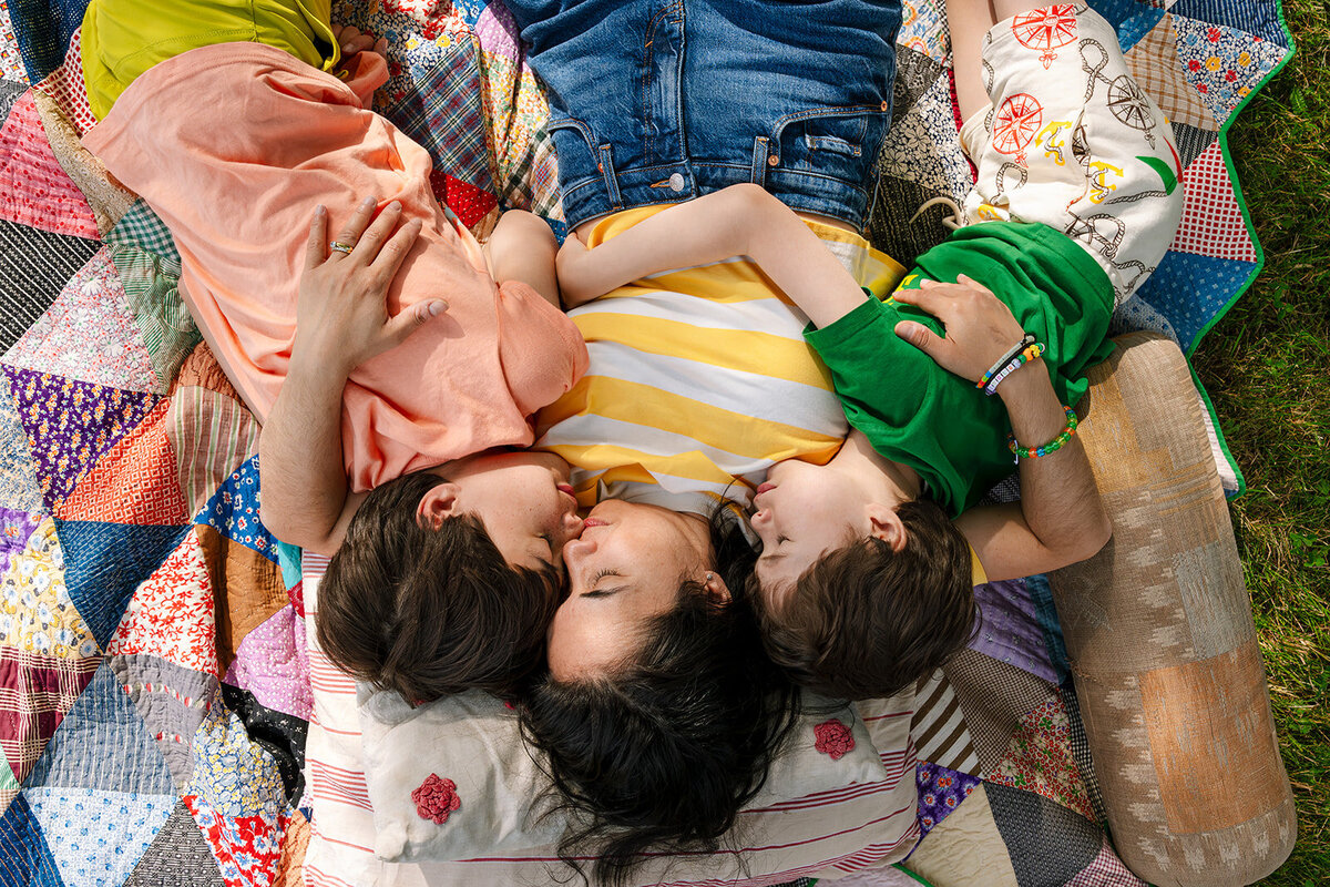 A close-up of a woman and child kneeling outdoors, with the woman holding a handful of small seashells and stones. The child holds binoculars and wears neutral tones, while the woman wears a colorful floral dress.