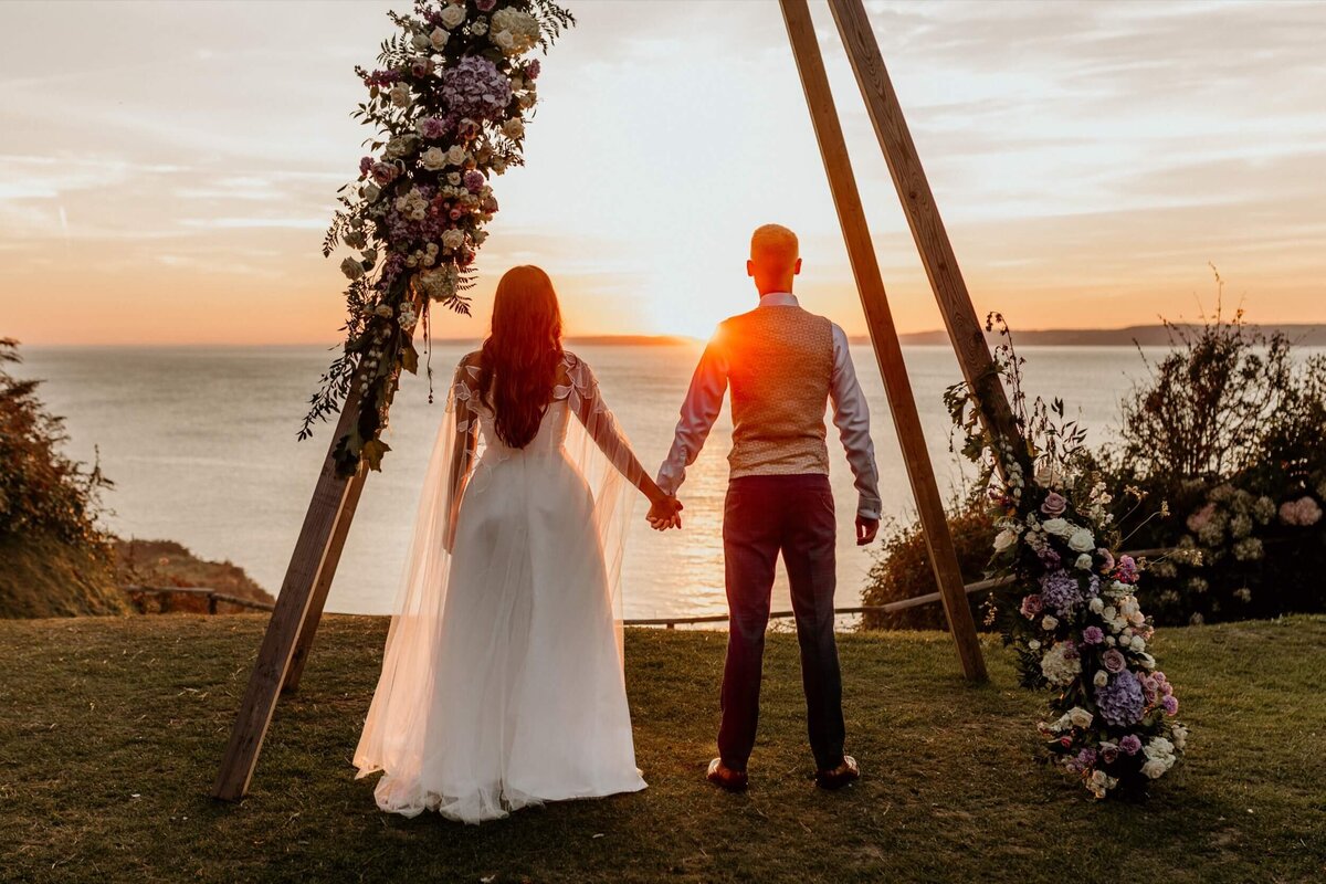 A couple standing under a wedding arch on the grass with a sunset in the background