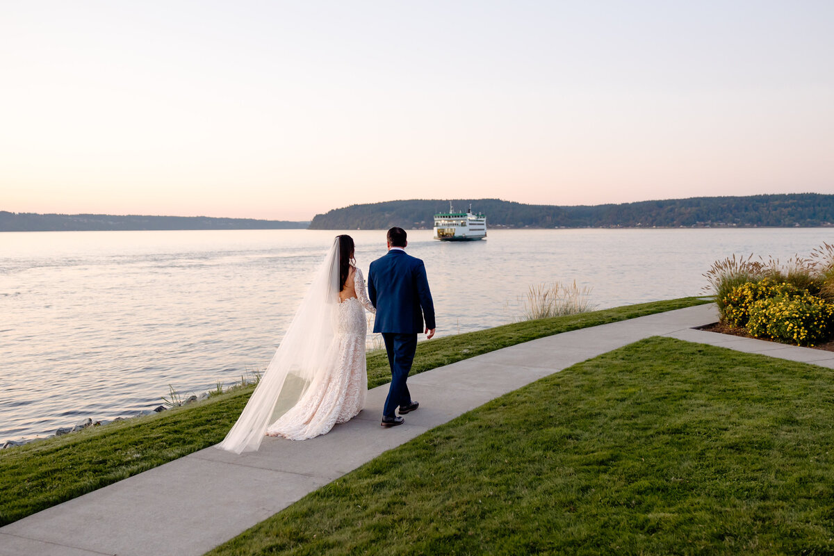 bride and groom walk along the waterfront at  tacoma yacht club with a ferry in the distance