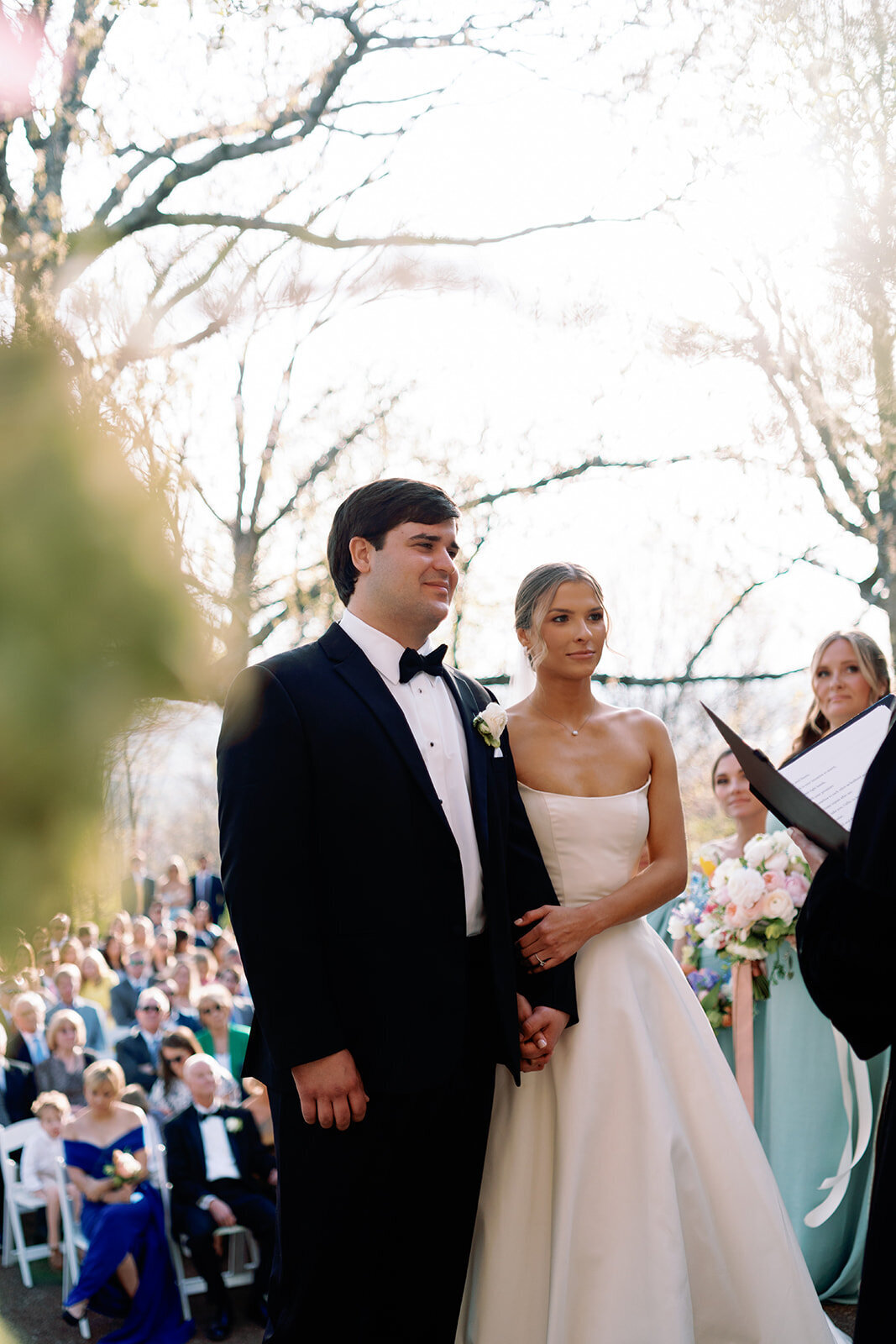 Bride and groom standing together and holding hands while listening to the officiant during their outdoor wedding ceremony at Burritt on the Mountain in Huntsville, Alabama, photographed by Perry Hancock.