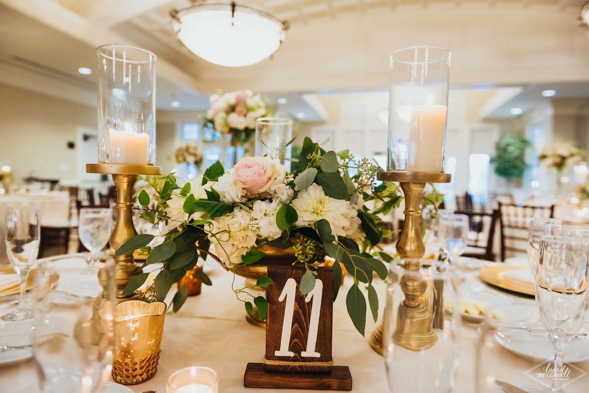 centerpiece of peach and ivory flowers with greenery, wooden table number sign, gold candle holders,and gold mercury votive candles