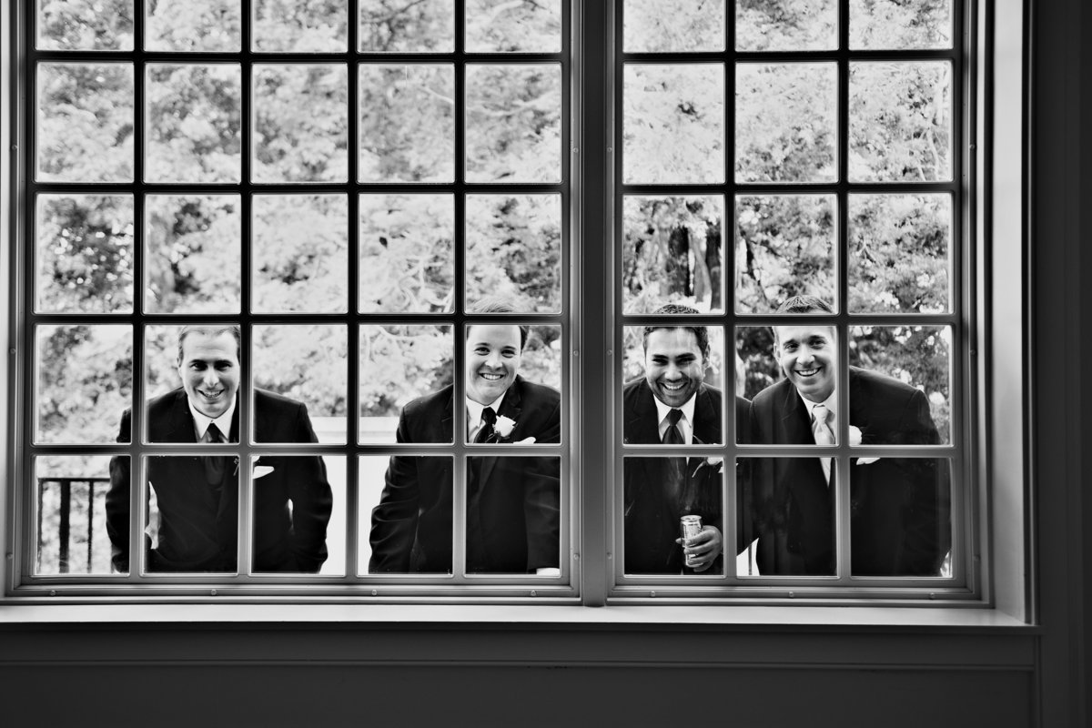 Groomsmen look through the window at wedding guests before the ceremony at Radnor Valley Country Club