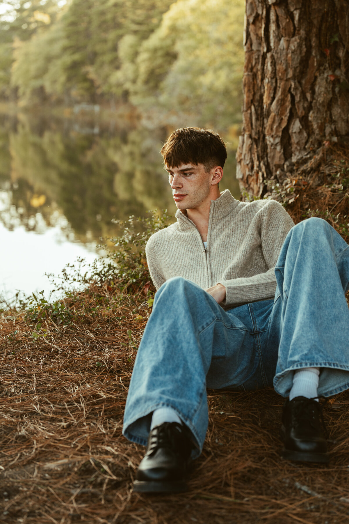 Young man sitting beneath a tree by the water—casual editorial portrait with soft golden light and relaxed, cinematic styling.