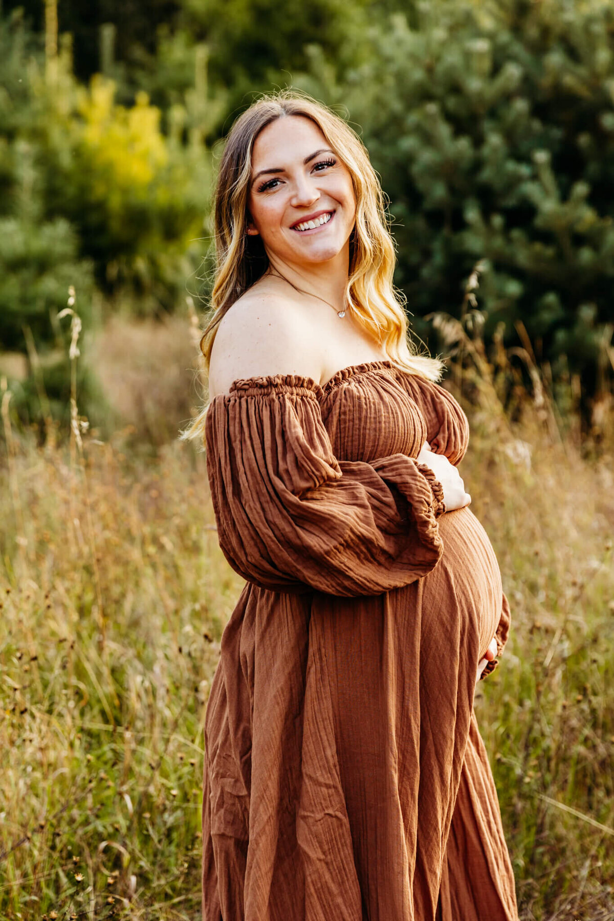 pretty mom to be in a brown flowy gown looking over her shoulder and embracing her baby bump  in a field near De Pere, WI.