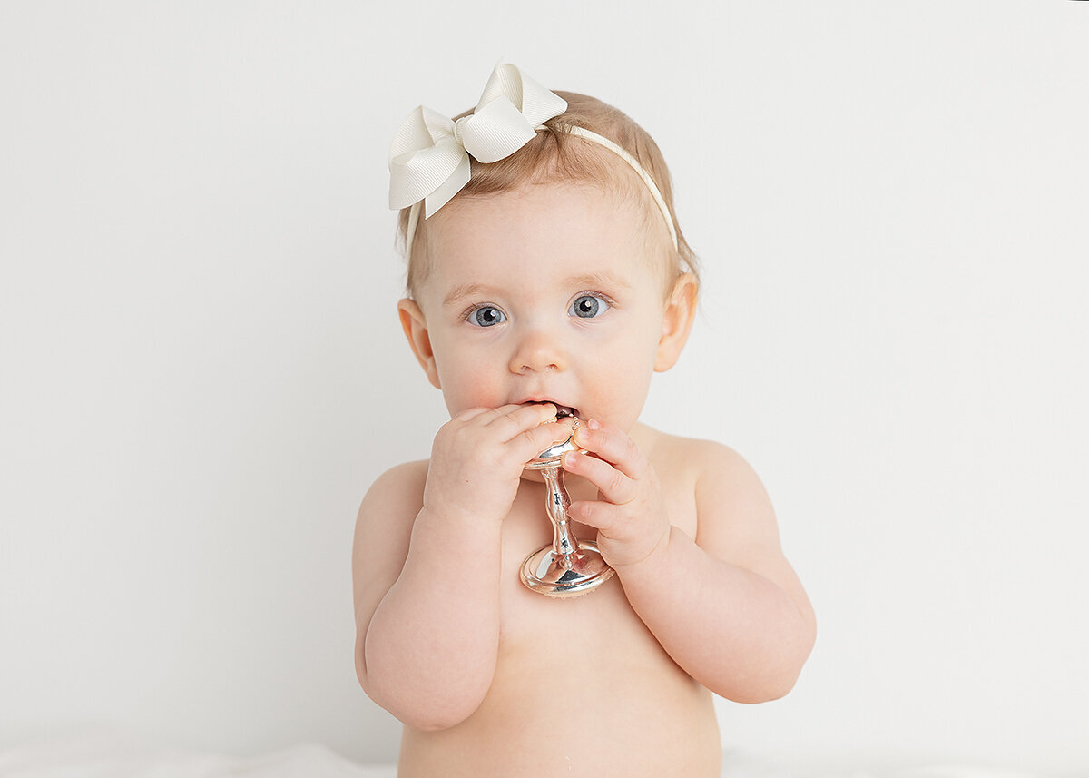 9 month old baby girl photographed in studio with a silver rattle and cream bow on her head