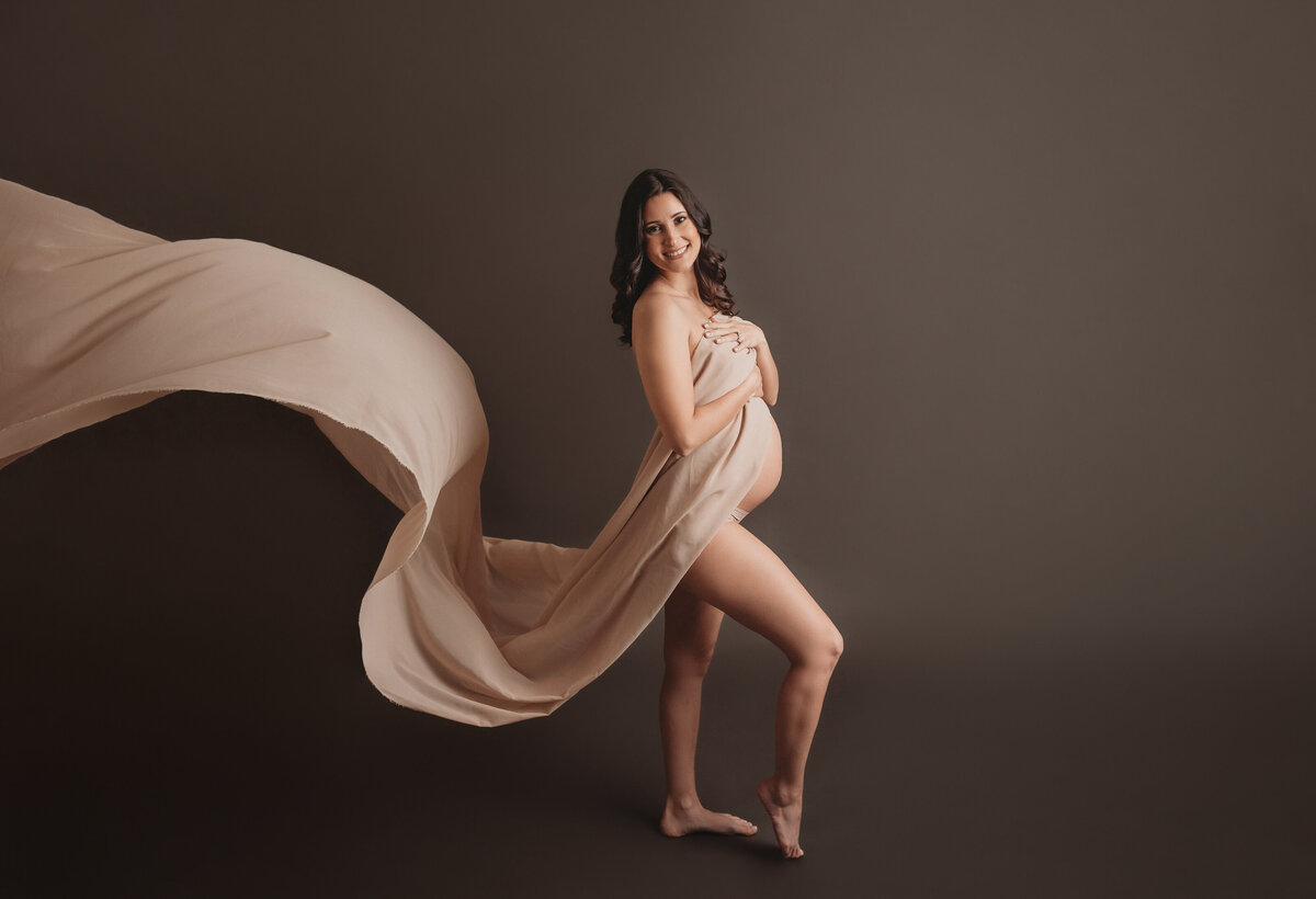Maternity portrait on dark gray backdrop of woman standing up with right leg  popped up while she is draped in light color chiffon fabric flowing behind her
