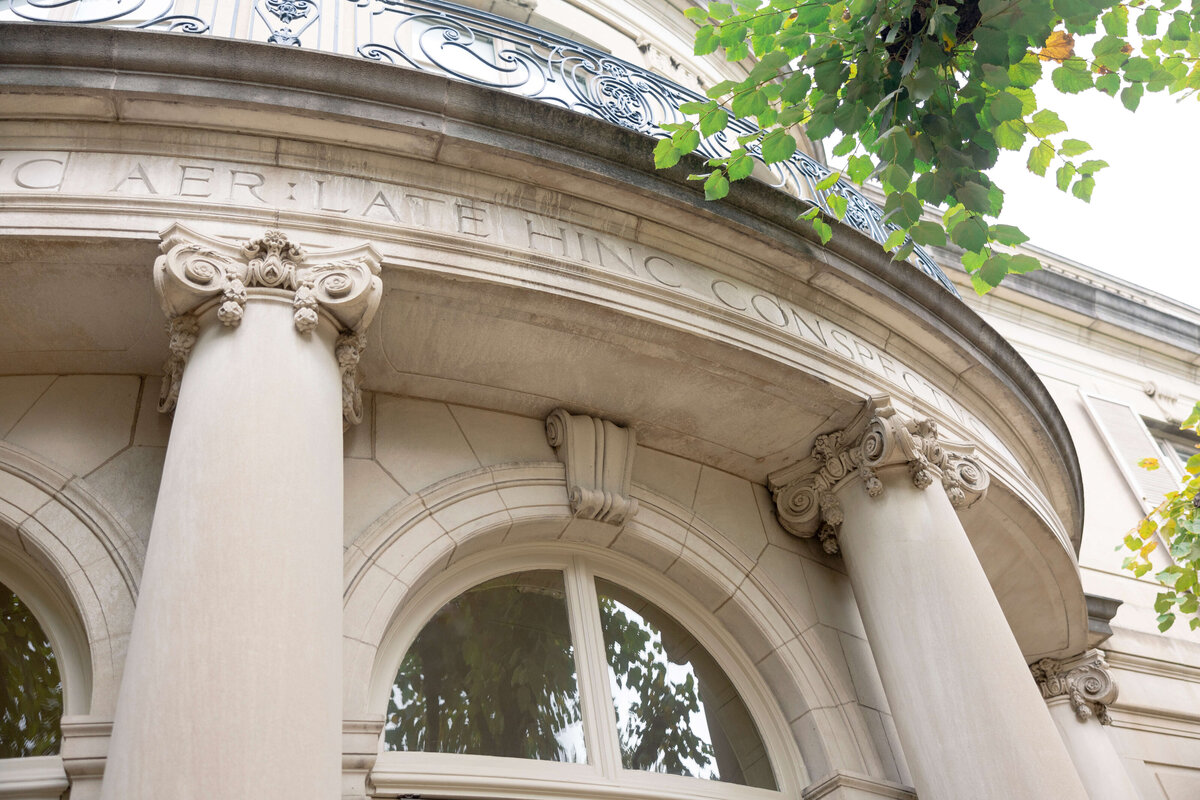 A close-up of a neoclassical building facade with four ornate columns and decorative moldings. Above the arched windows, theres an inscription on the stone. Green leaves partially frame the top right corner of the image.