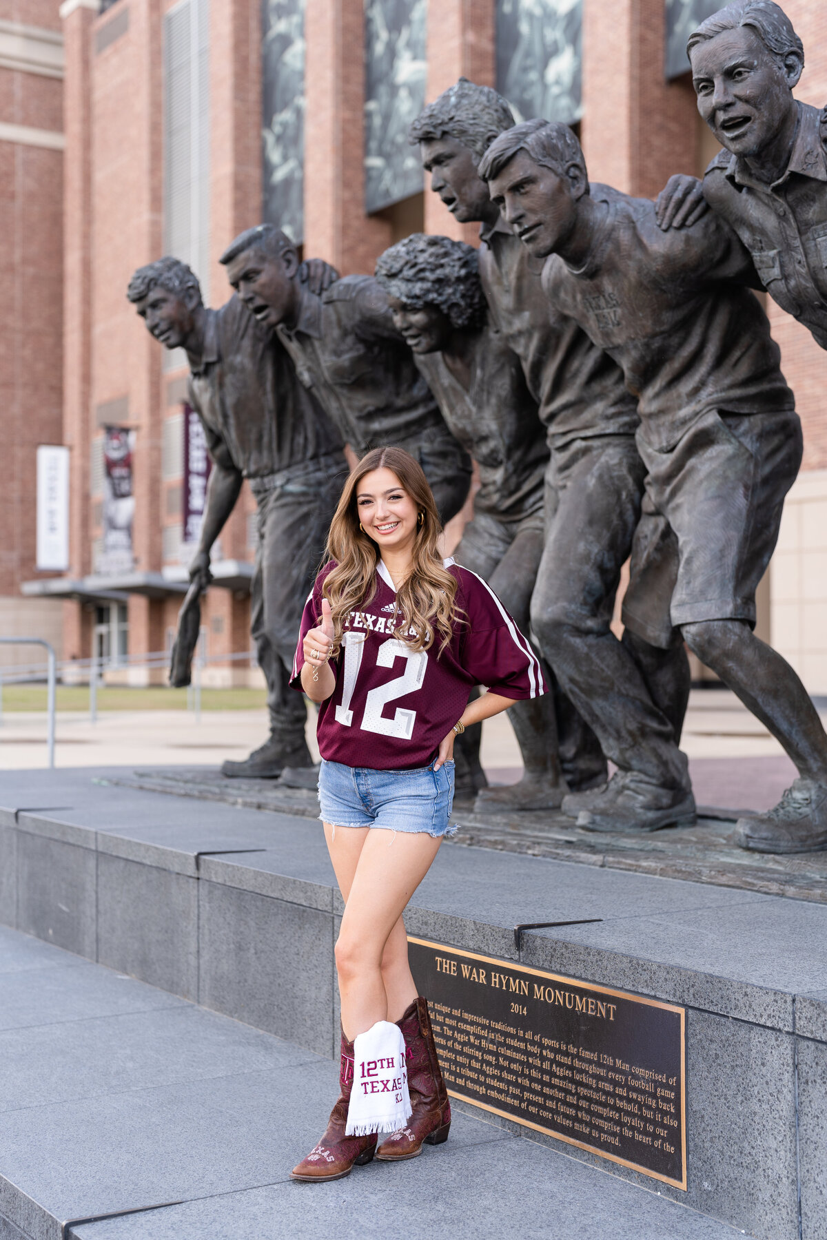 Texas A&M senior girl in maroon jersey and cowboy boots with thumbs up smiling in front of War Hymn statue at Kyle Field
