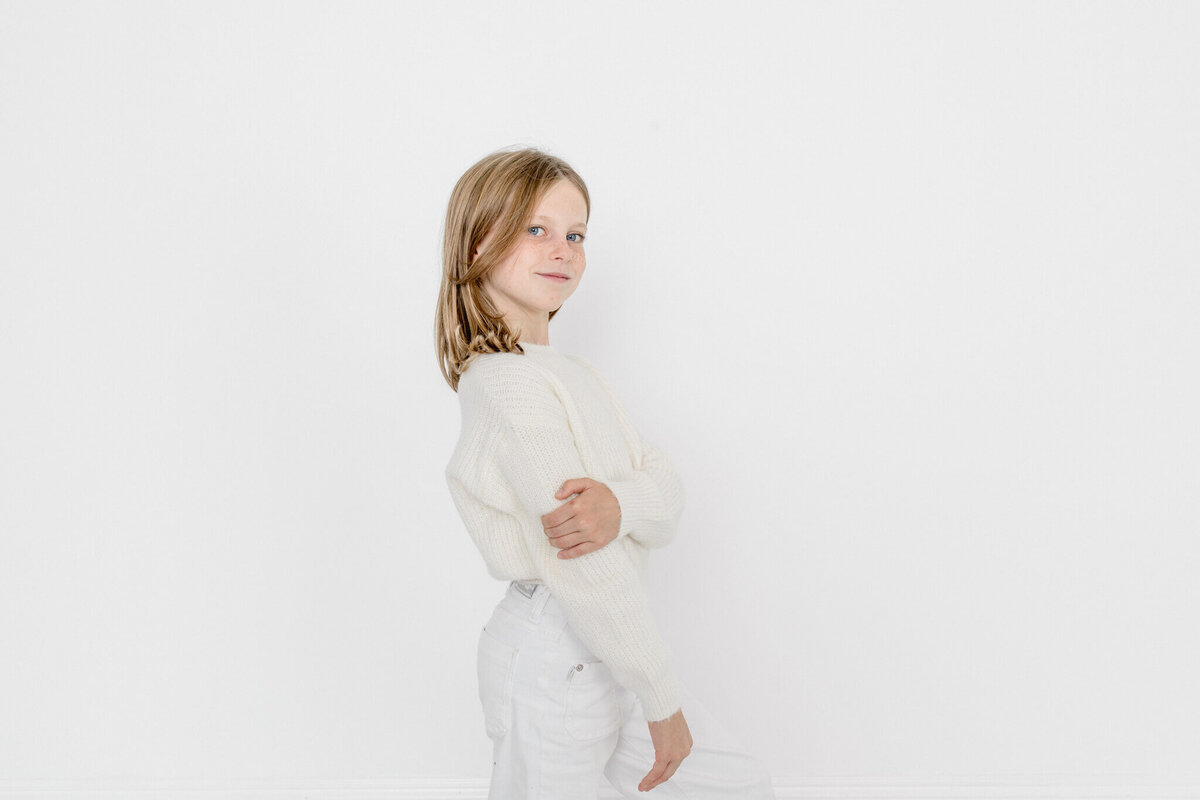 A pre-teen girl with shoulder length blonde hair wearing all white, facing sideways from the camera, looking at the camera and smiling very subtly and holding her arm with one hand in a white studio in Tustin.