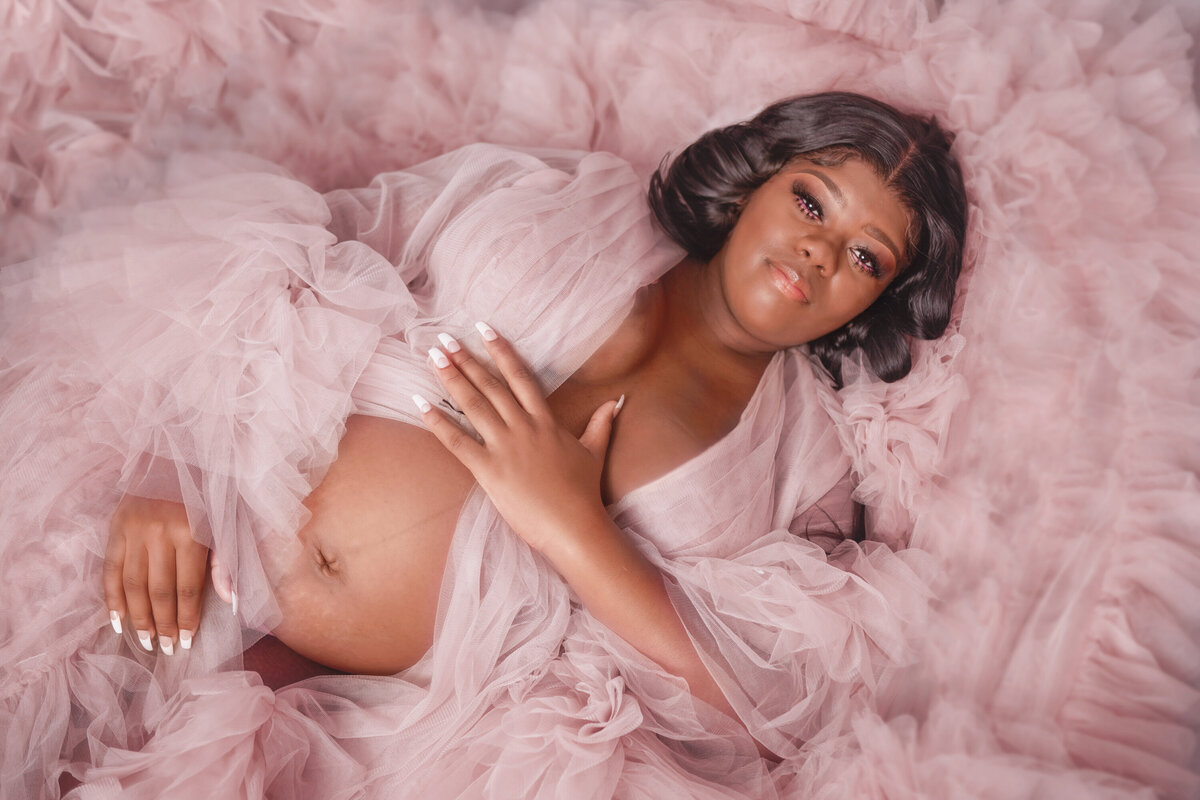 Black woman in a pink tulle robe laying on the floor.  Her pregnant belly is exposed and she is holding her robe closed.  She has black hair and long eyelashes.