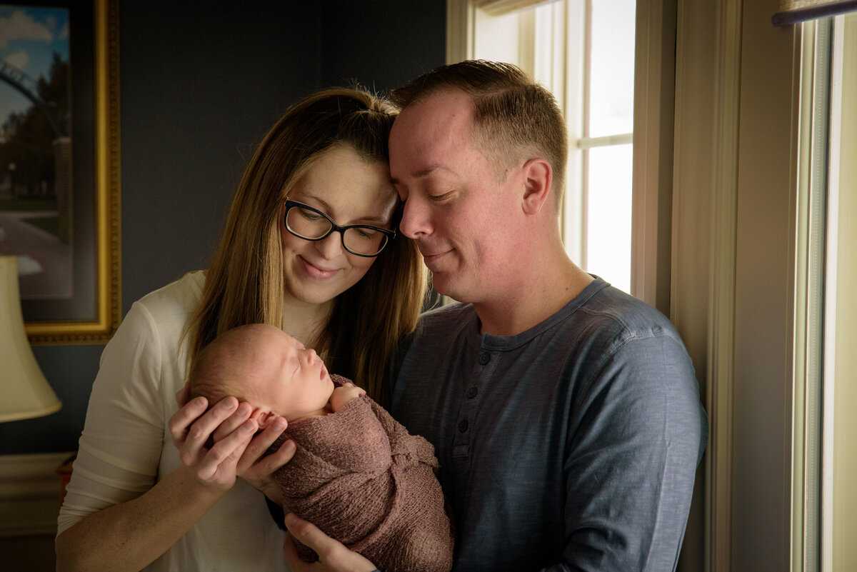 Mom & Dad are holding and adoring their new baby girl who is wearing a dusty rose wrap in front of a window in their home in Green Bay, Wisconsin.