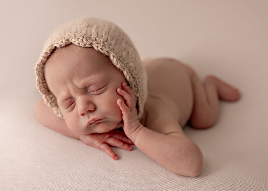 newborn boy posed chin on hands with a knit bonnet.  Cream backdrop