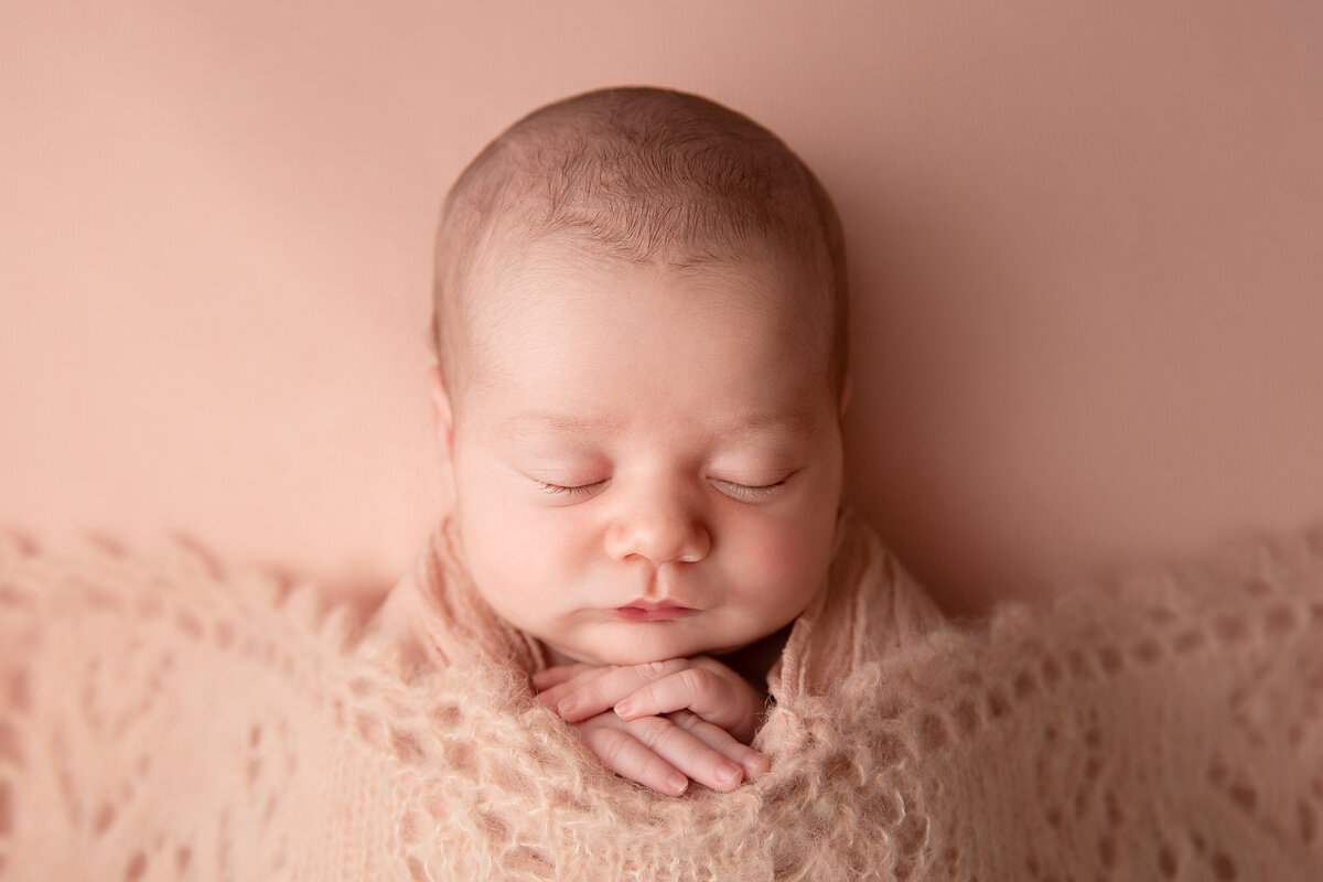 Newborn baby girl sleeping wrapped in pink with a pink knitted blanket over her.