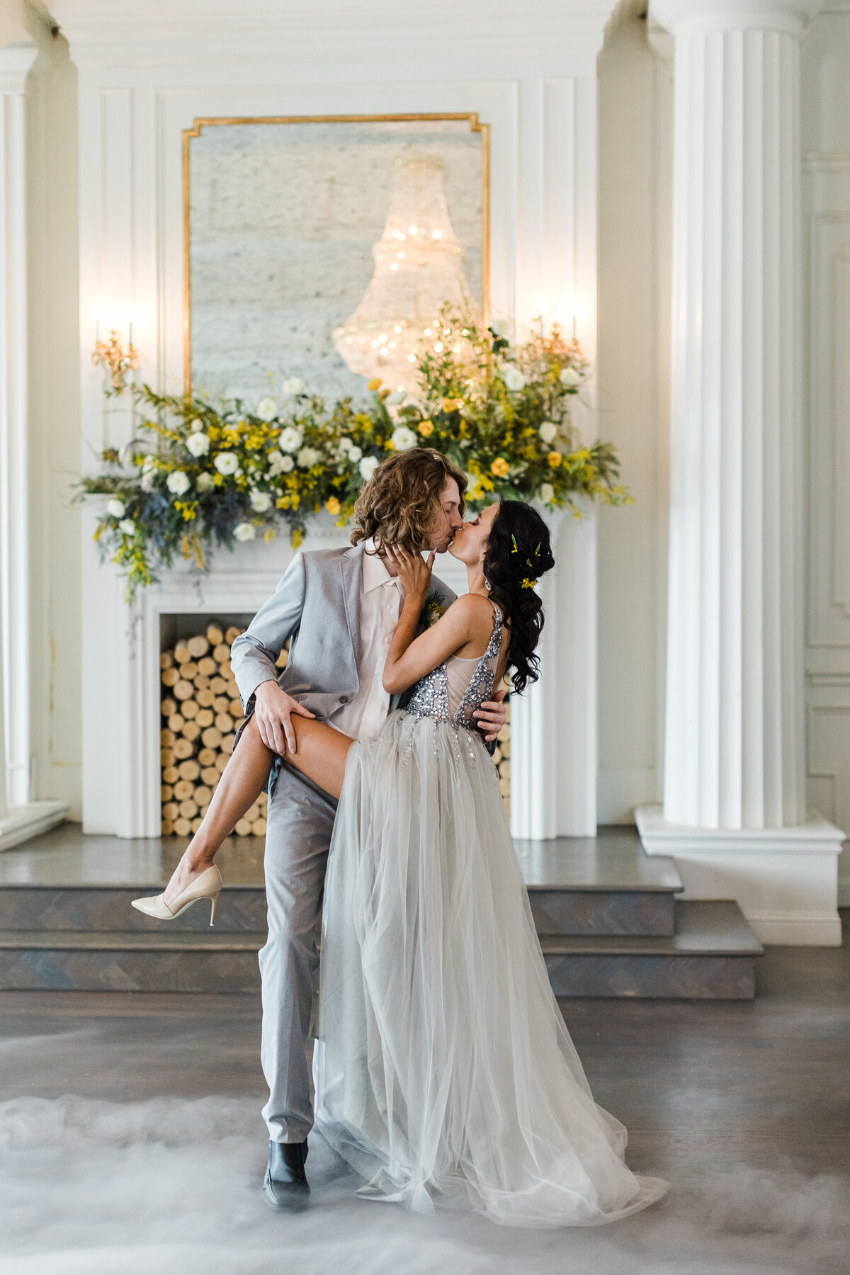 A portrait of a bride and groom sharing a passionate kiss at The Mason in Dallas, Texas. The bride is on the right and is wearing a sparkly, silver dress. The groom is on the left and is wearing a grey suit with a boutonniere. They are backed by a large, fancy fireplace adorned with a large floral arrangement.