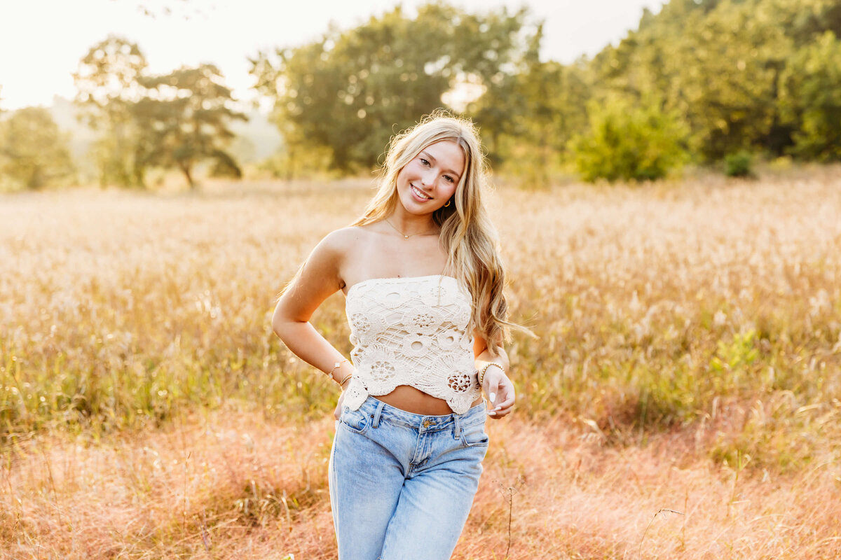 gorgeous high school girl standing in a glowing field at sunset with one hand in her back pocket and smiling.
