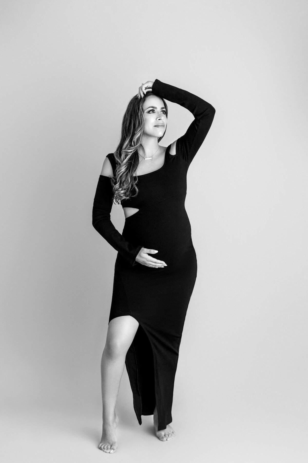 A black-and-white maternity portrait of a pregnant woman standing barefoot against a light gray background. She wears a stylish, form-fitting black dress with cut-out shoulder details and a thigh-high slit. One hand cradles her baby bump, while the other is raised above her head, gently resting on her hair. Her expression is confident and serene, capturing the elegance and strength of pregnancy in a minimalist setting.