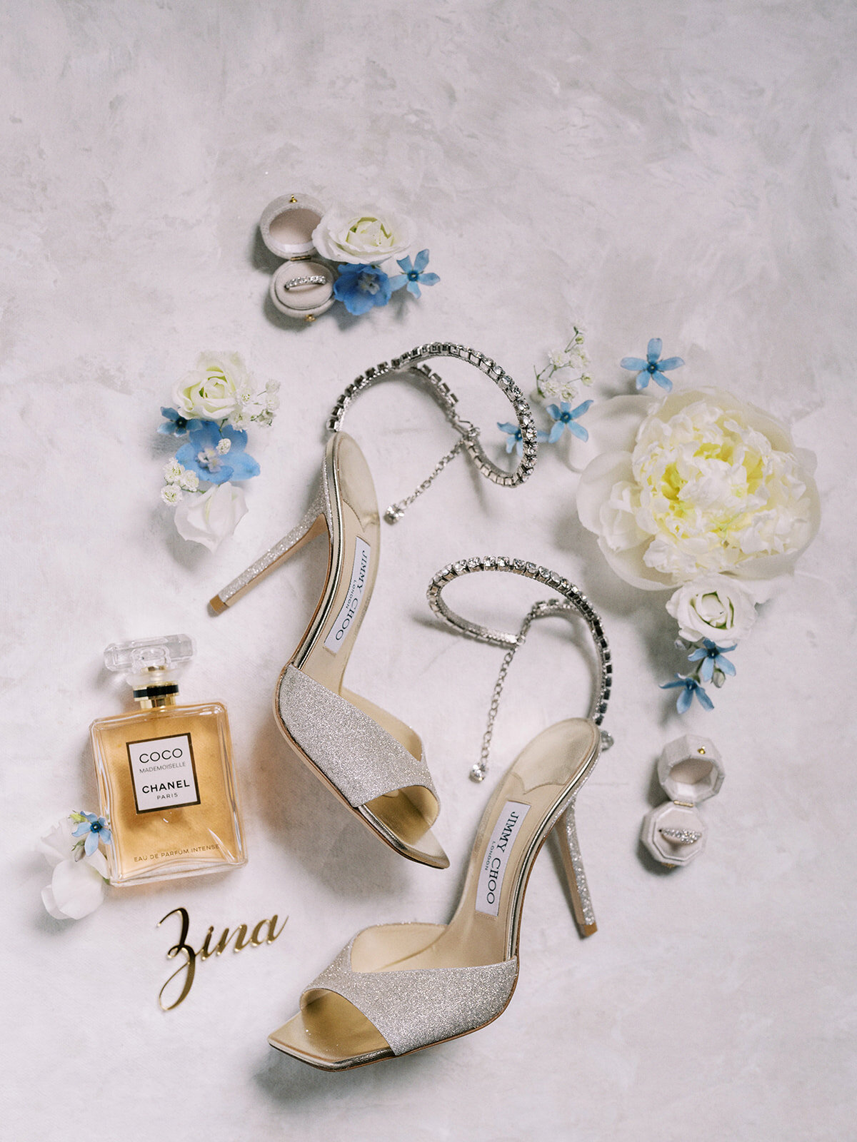 Pair of silver high-heeled shoes, a bottle of Chanel Coco perfume, and floral decorations arranged on a textured surface, with the word "Zina" in gold—a perfect setting for a destination wedding in Canada.