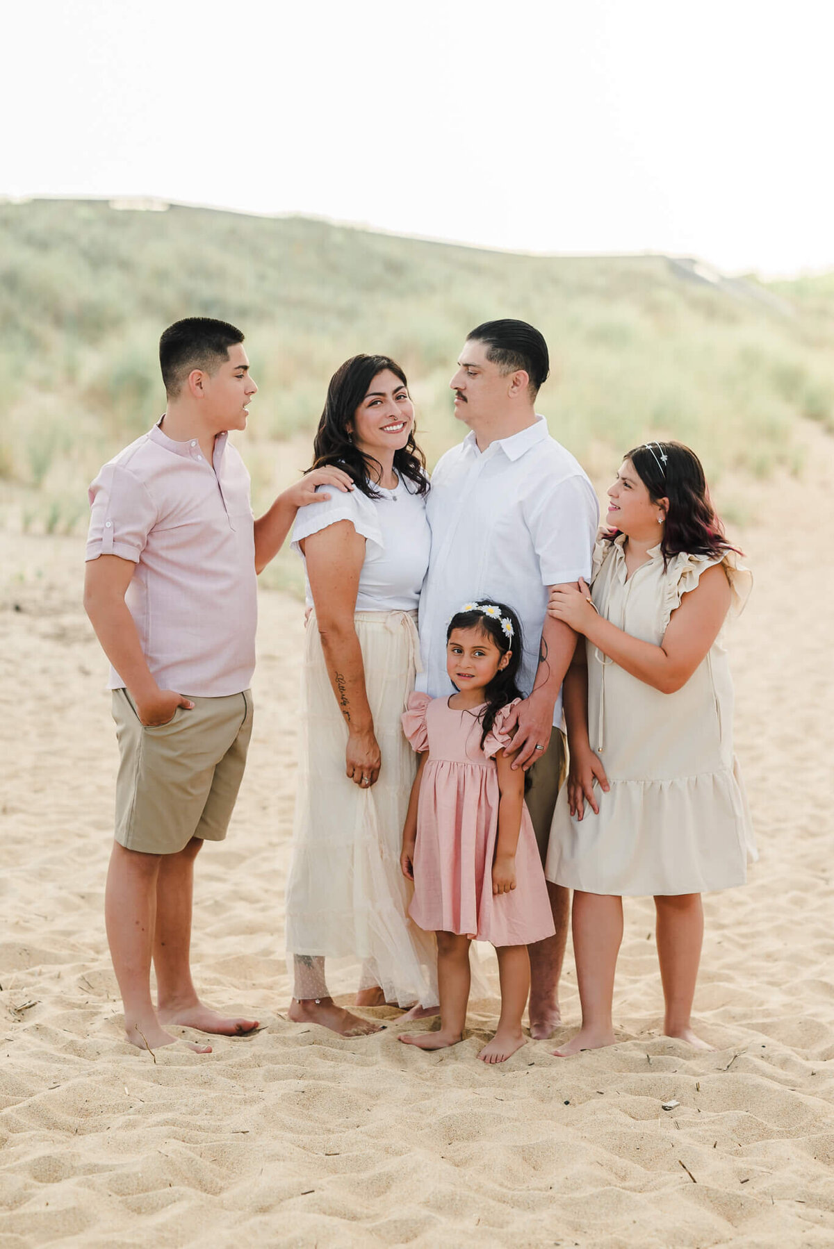 A family of five poses in front of the dunes at a beach in the Outer Banks. The dad, teenage son, and oldest daughter are looking at the mom. The youngest daughter smiles for the camera.