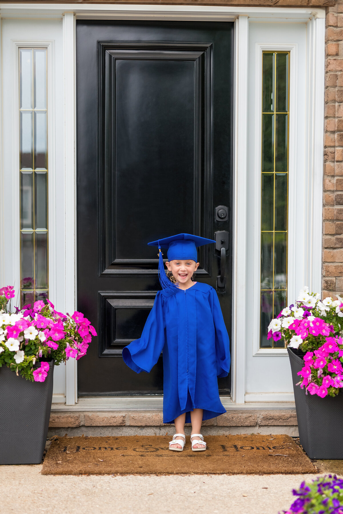 Graduation milestone of a young girl in a blue cap and gown smiling at her front door—beaming with pride and promise.