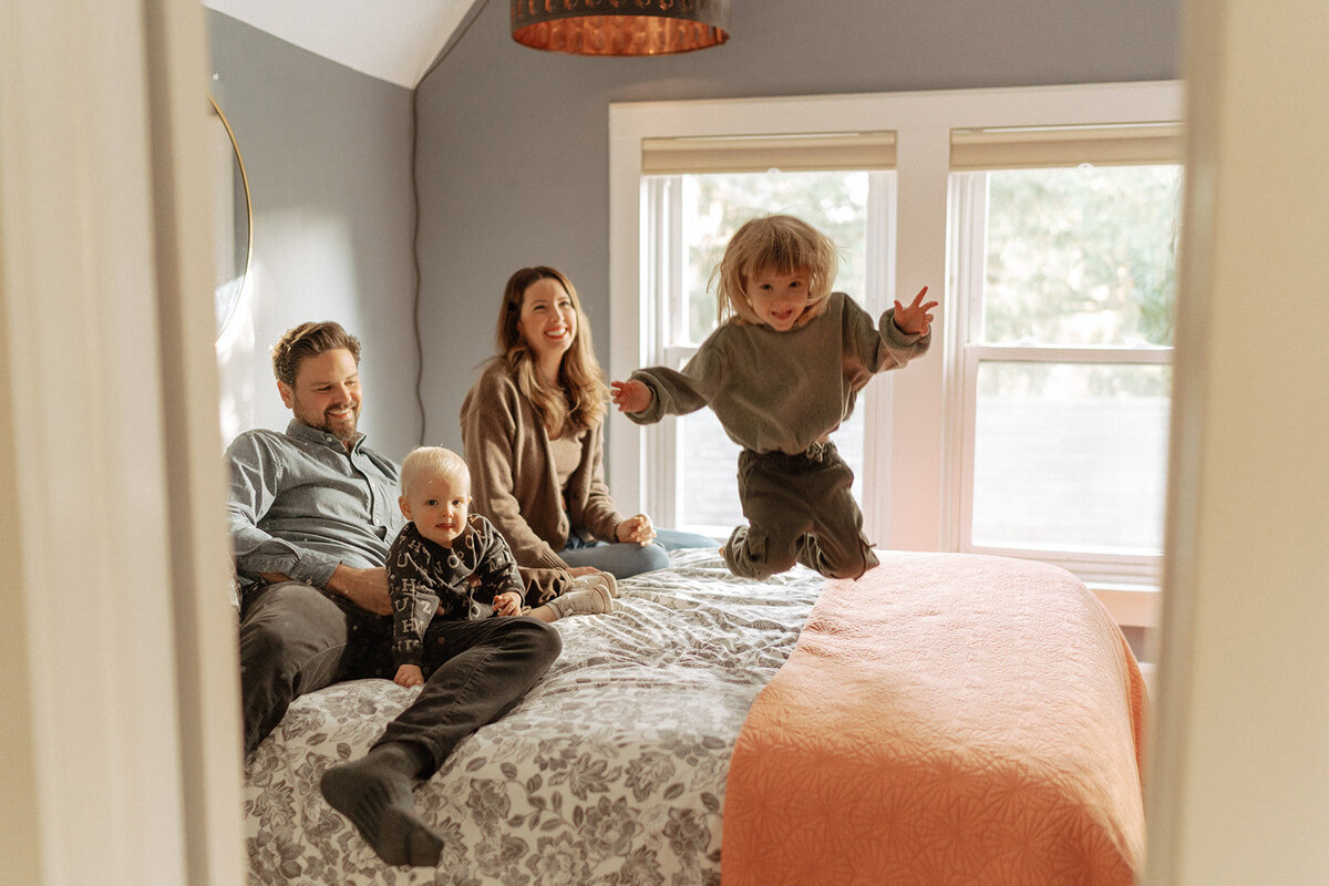 couple sitting on bed hold a baby while a toddler jumps off during family photos captured by NYC family photographer Elsie Goodman 