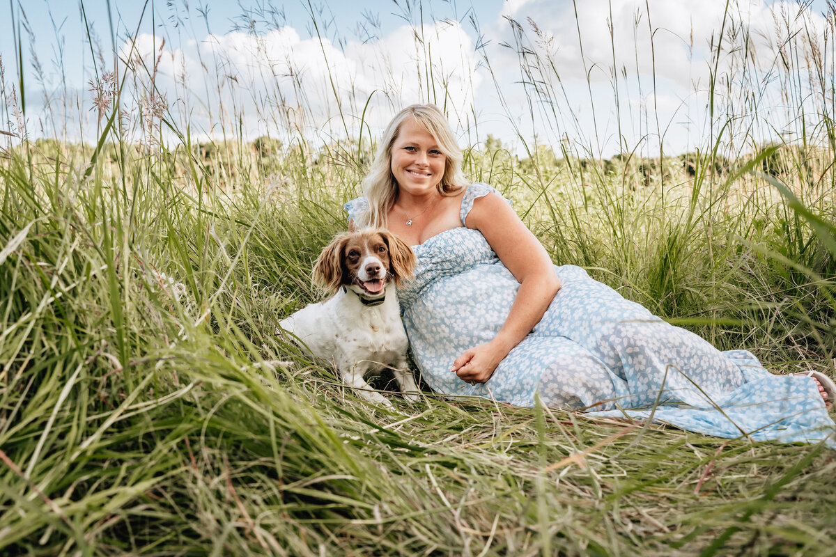 A pregnant woman in a light blue floral dress sits in a grassy field with a dog by her side, both looking content and relaxed.