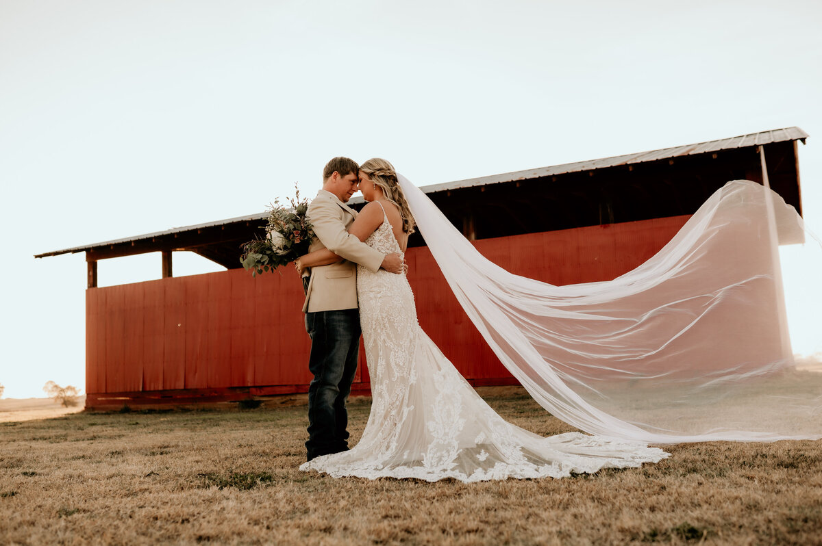 bride and groom embrace each other while they hold their hips and place their foreheads together with a red barn in the background while the brides veil blows in the wind