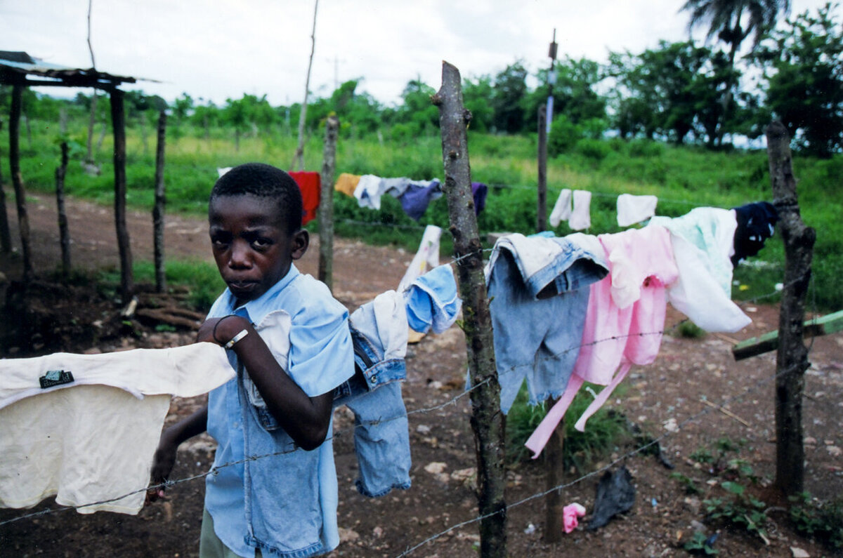 Boy leans on barbed wire fence with laundry drying for portrait in Haitian Batey.