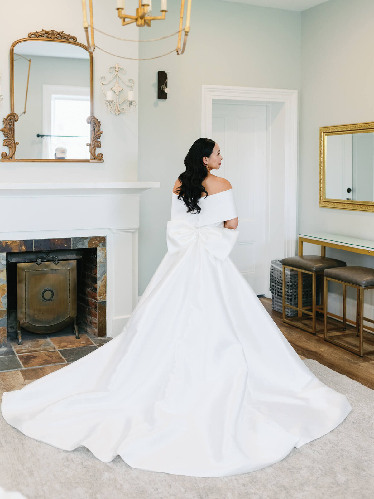 A bride with long, dark hair stands in an elegant room, wearing an off-the-shoulder white gown with a large bow.