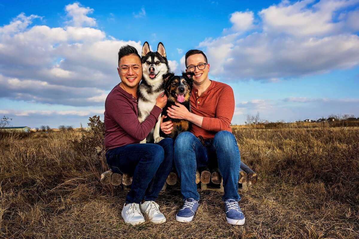 LGBTQ+ couple wrap their arms around their Husky and Australian Shepherd dogs, all of them sitting closely in a hay field.