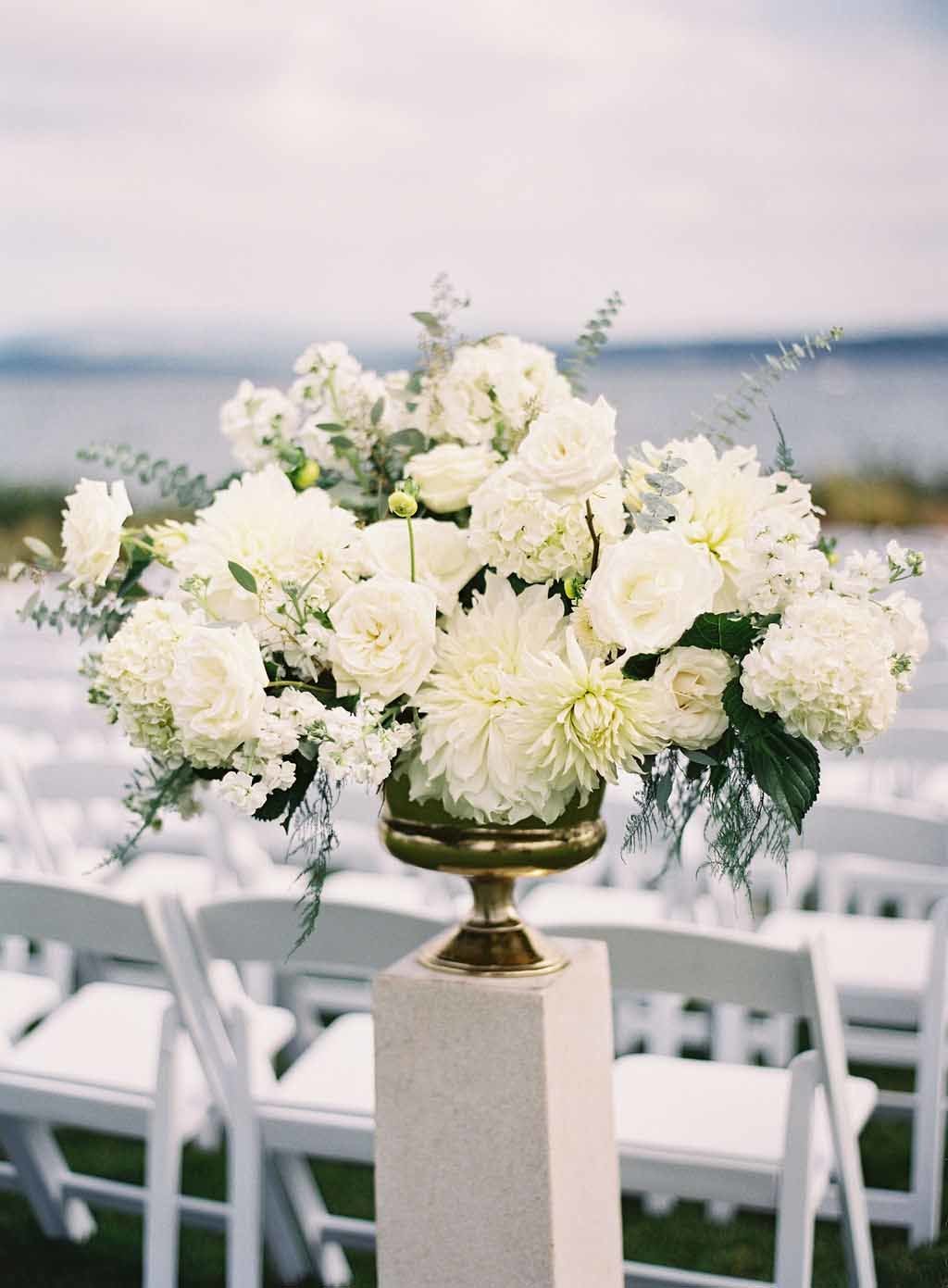 large white flower arrangement in gold urn on top of white stone pedestal for wedding ceremony