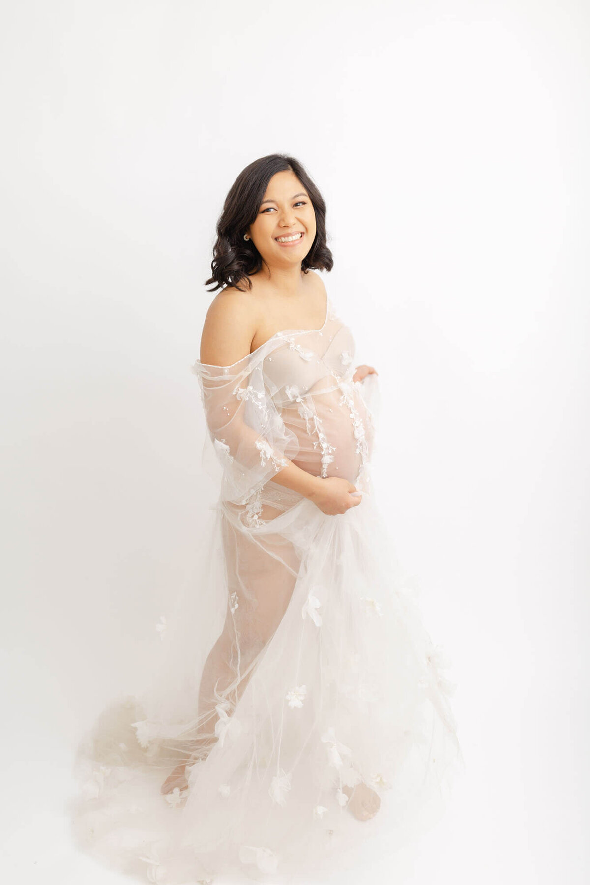 Pregnant Woman in see-through tulle dress with beading work on it. She is swaying her dress a bit and smiling at the camera. She has shoulder-length black hair in loose curls and professional hair and makeup done for her maternity portrait session in an all-white photography studio in Portland, OR. Photo by Ashlie Behm Photography