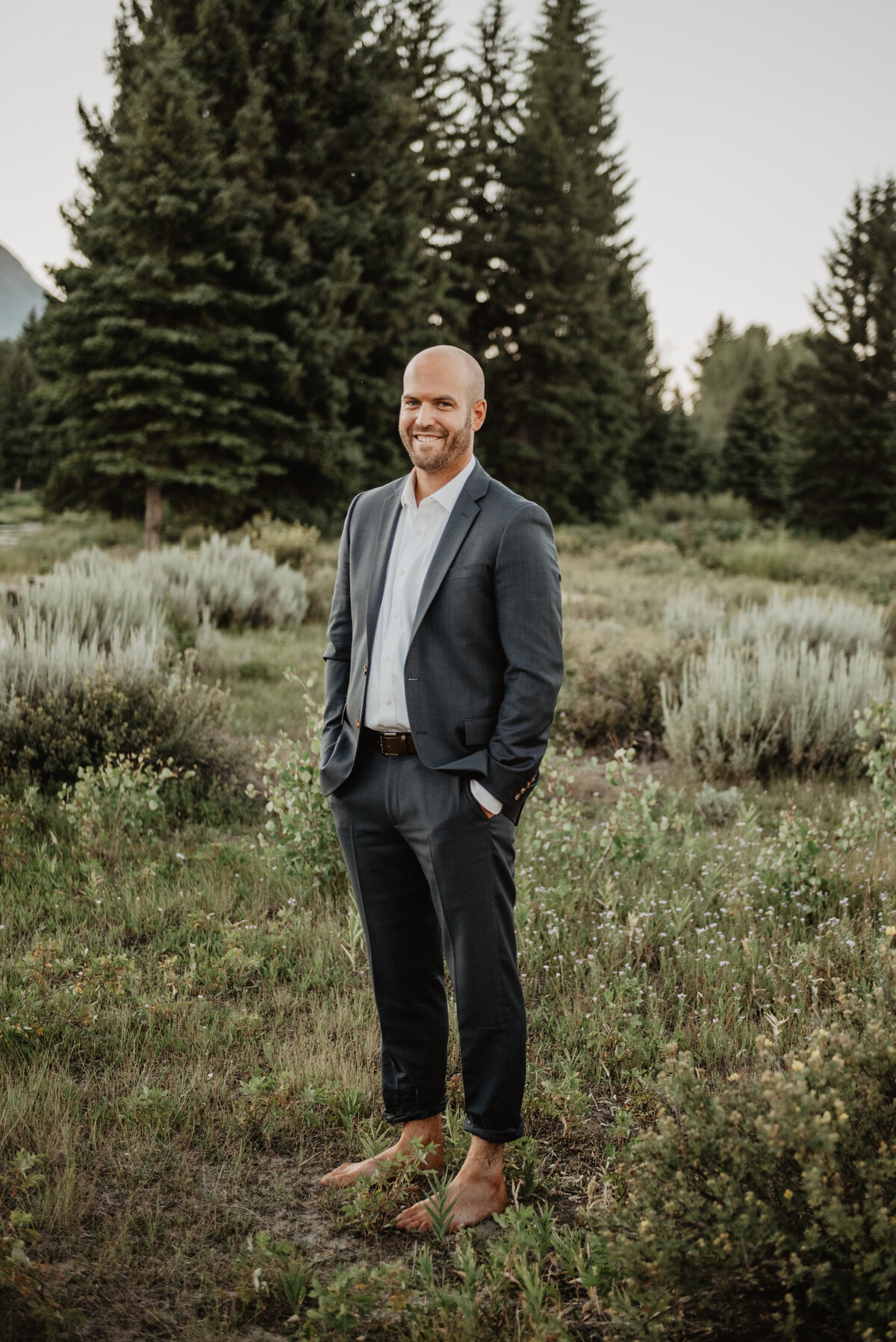 jackson wyoming photographer captures groom standing in a field wearing his wedding day suit with his hands in his packets smiling
