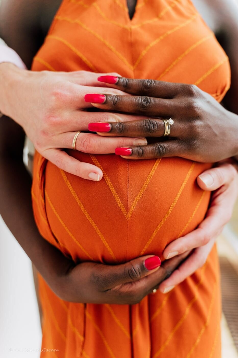 Close-up of a pregnant woman wearing an orange dress with bold diagonal stripes, her belly embraced by her partner’s hands and her own, all hands resting together with red-painted nails and wedding rings.