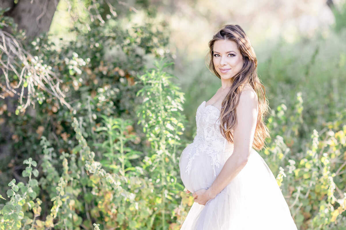 A pregnant woman in a flowing white dress stands outdoors in a lush, green setting, holding her belly with both hands and smiling softly at the camera. Sunlight filters through the leaves, creating a warm, serene atmosphere.