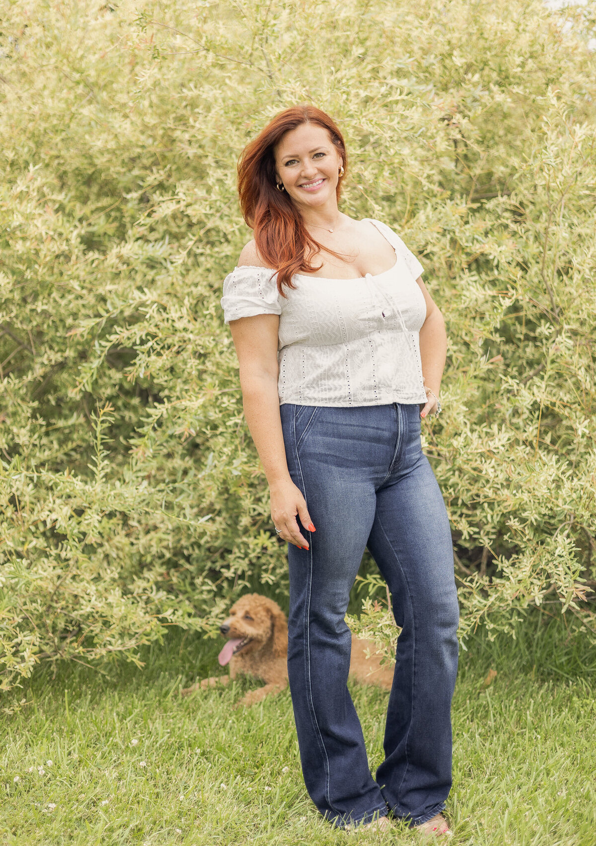Mom is standing in the grass in front of the bush with their light brown dog laying in the grass behind her.  Mom is smiling at the camera.  Mom is wearing blue jeans, short sleeve white shirt, and has long red hair. 