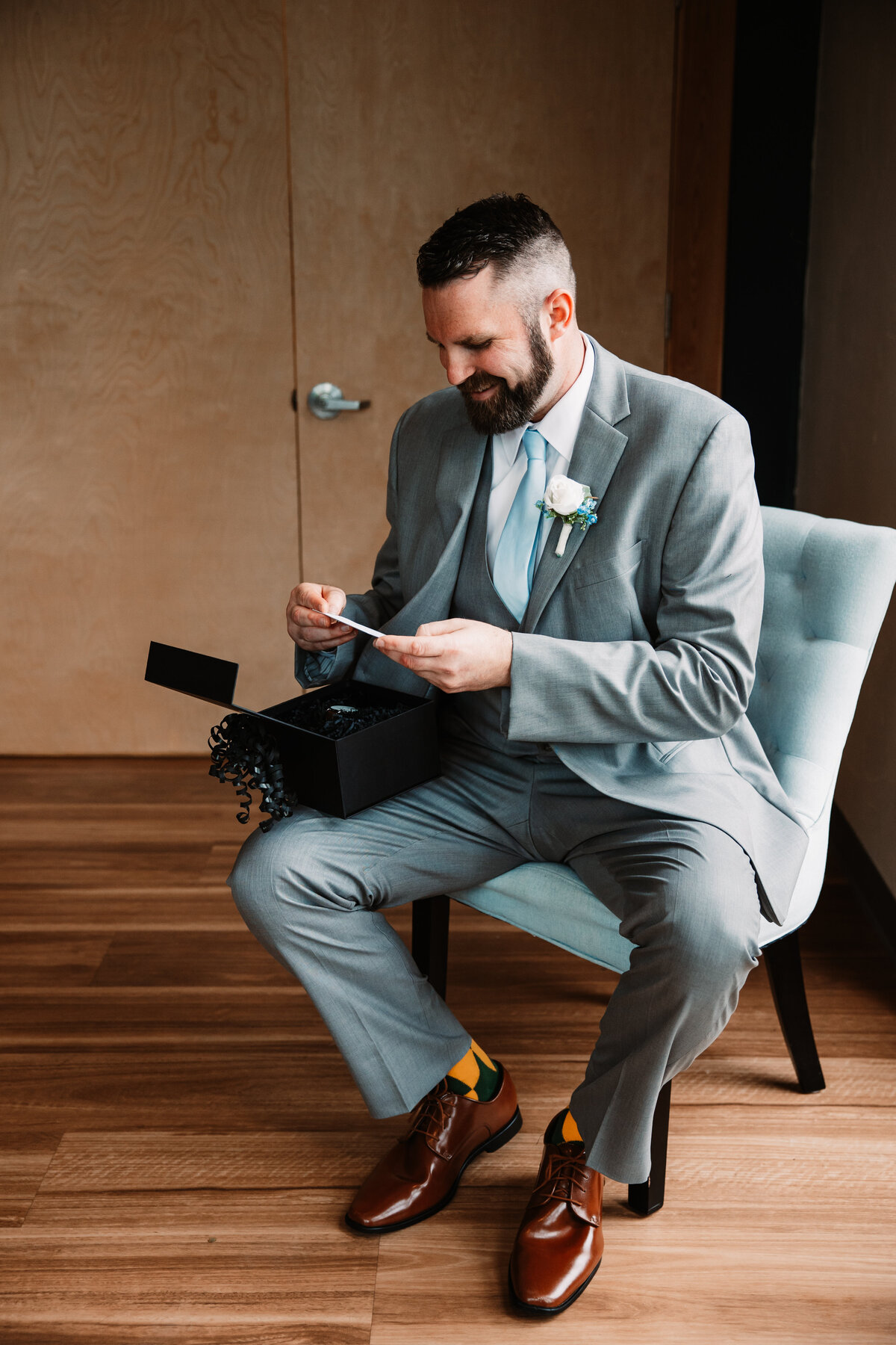 A groom in a grey suit sits in a chair reading a card and opening a present under a window