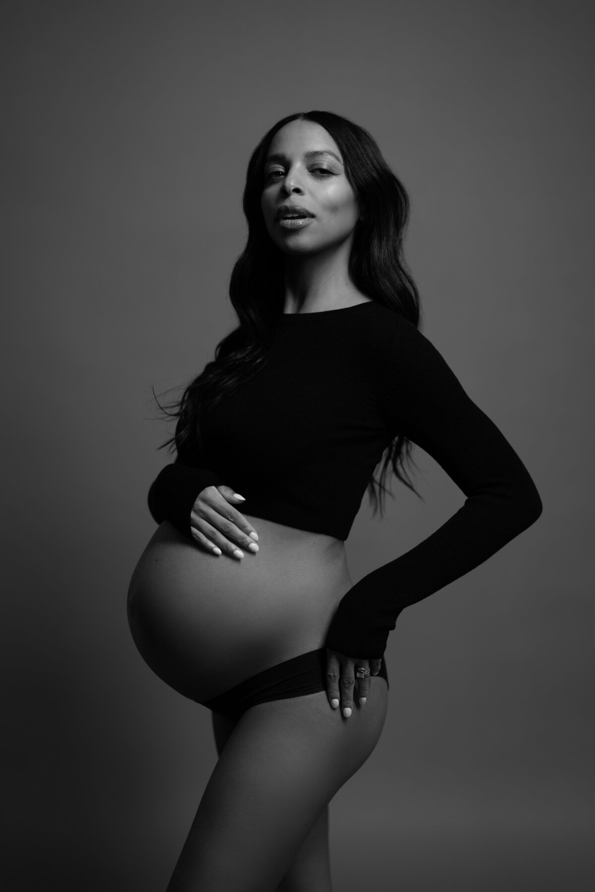A confident studio maternity portrait showcasing the expectant mother in a fitted black top with her hand on her hip, taken in NYC.