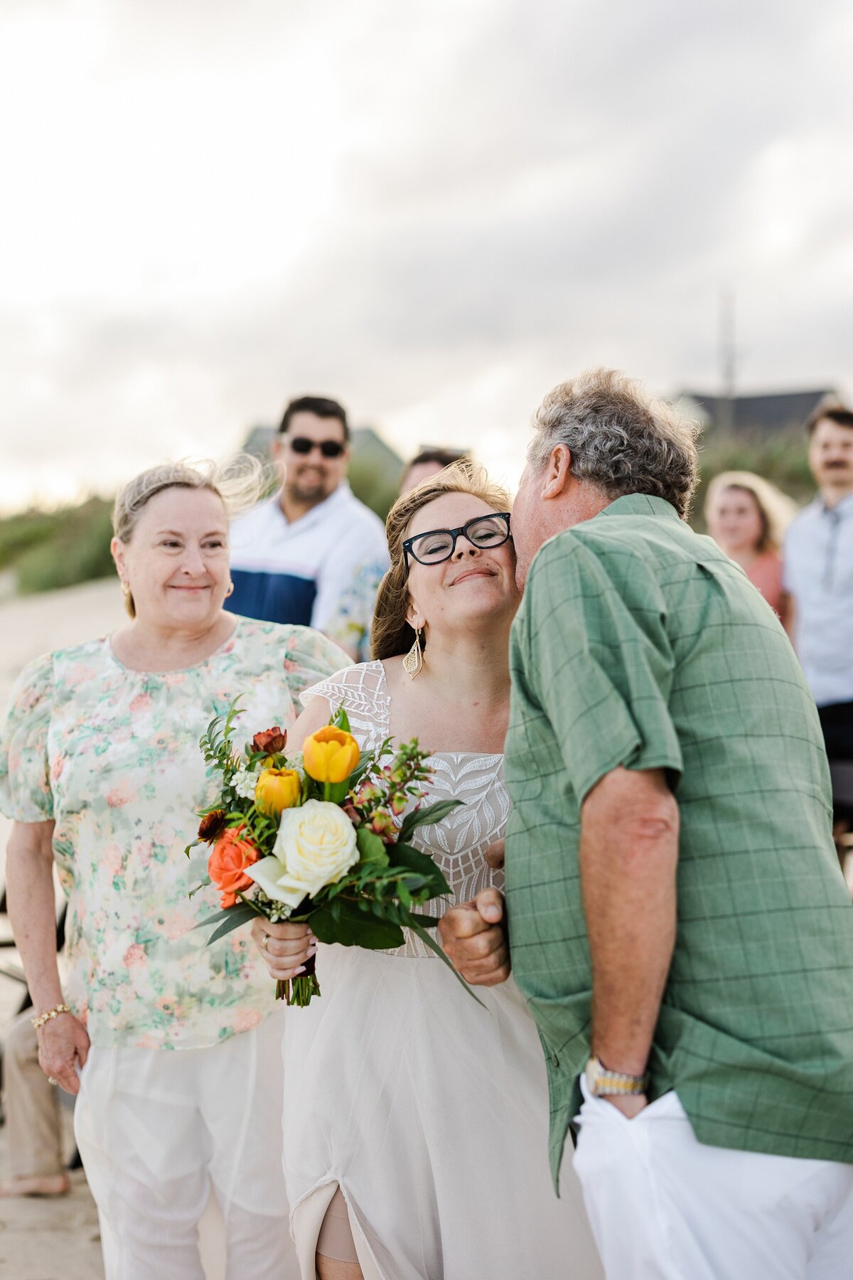 Candid shot of a bride receiving a kiss on the cheek from her father as her parents finish walking down the aisle during her wedding ceremony at Crystal Beach, Texas. The bride is wearing a detailed white dress and is holding a bouquet. Guests can be seen in the background standing on the sand.