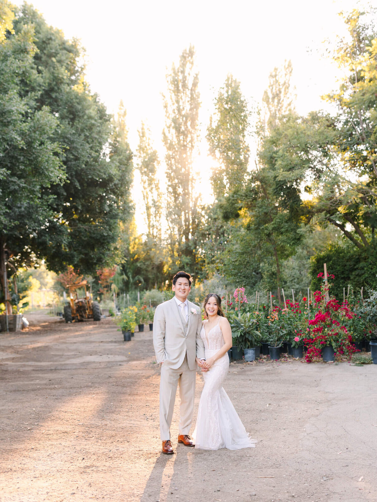 A couple stands smiling on a sunlit path in a garden, surrounded by lush green trees and vibrant red flowers.