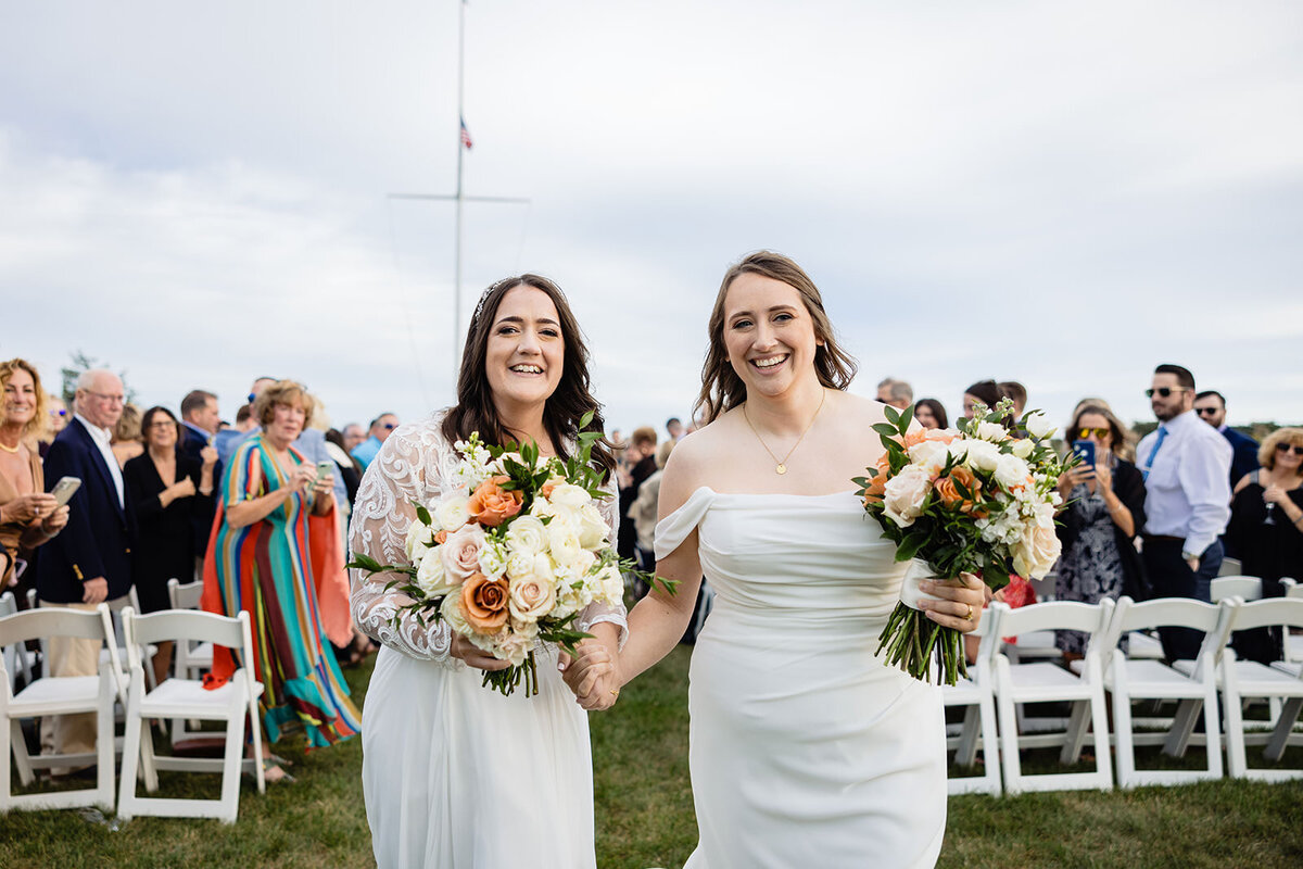 Two brides hand in hand, walking down the aisle, smiling broadly. They are both in white dresses and holding bouquets with orange and white flowers, with guests standing on either side.