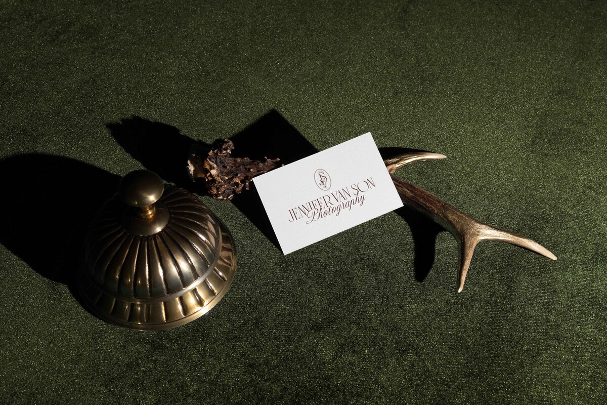 A business card reading "Jennifer Rawson Photography" is propped on a deer antler beside a bronze bell, all placed on a green textured surface—an inspiring scene for any showit website designer seeking creative branding ideas.