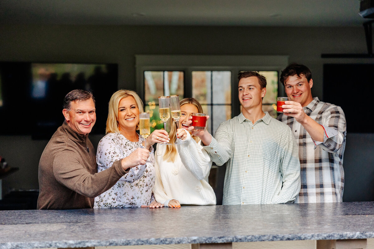 A group of five people stands behind a counter, smiling and raising glasses in a toast. The softly lit room captures their joy beautifully, reminiscent of a scene from Brainerd Lakes photography. Both men and women, dressed casually, exude warmth and camaraderie.
