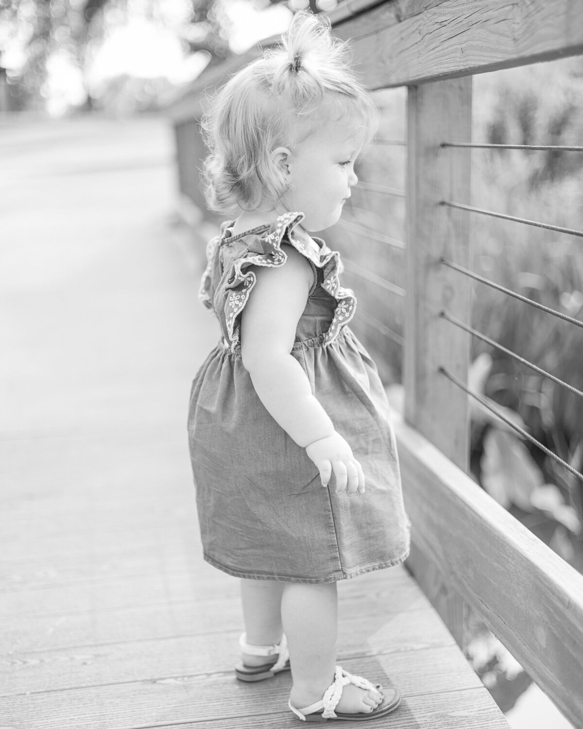 black and white photo of toddler girl on a bridge looking out over the pond, wearing a dress, and has pigtails