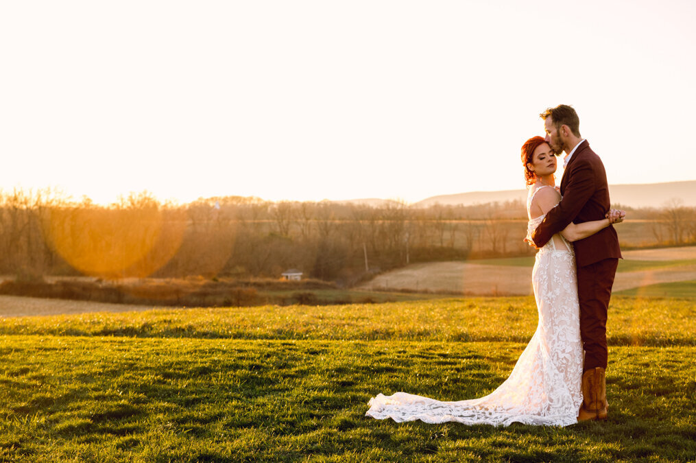 A bride and groom embrace in a field at sunset.