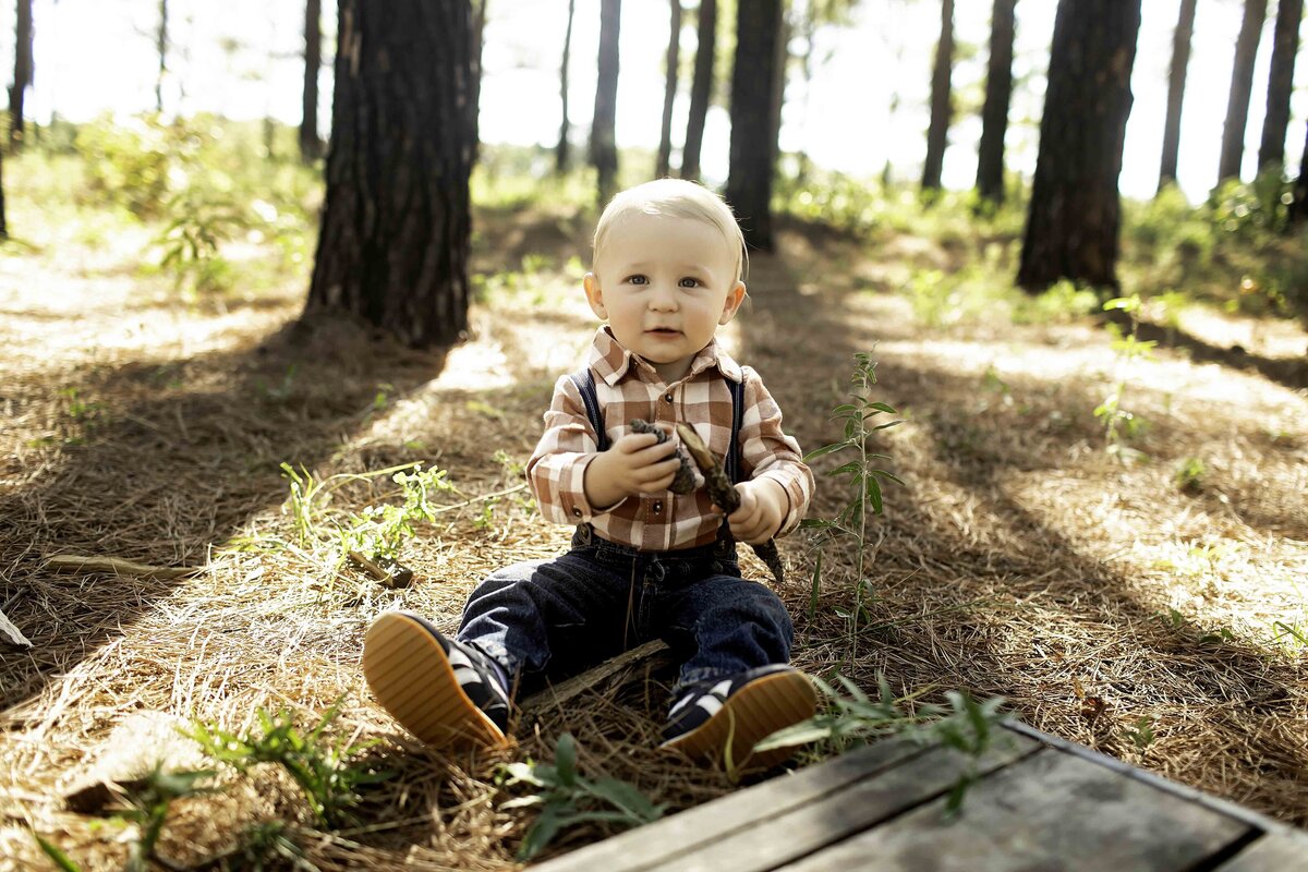 One-year-old birthday boy in suspenders and overalls, exploring the pine tree forest with excitement