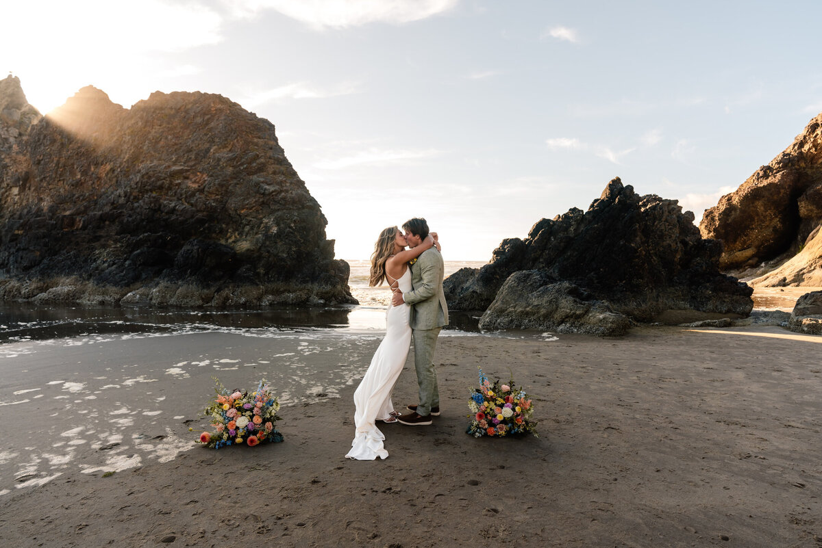 A bride and groom share their first kiss as married during their Oregon coast elopement. The waves inch in around the sea stacks they are composed between and the wind billows the brides dress.