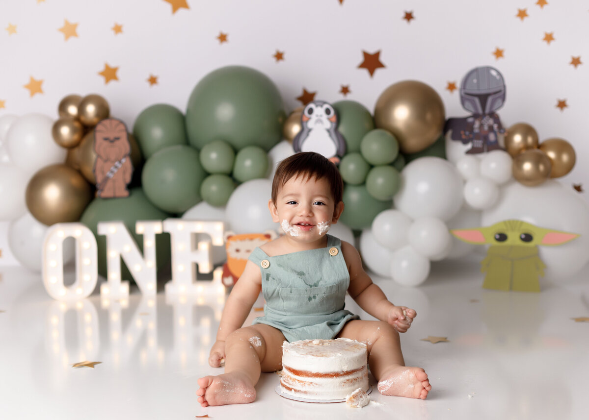 Star Wars themed cake smash in Wellington and West Palm Beach Florida portrait studio. Baby boy is wearing soft green overalls sitting behind a white naked cake. The background is cream with stars and sitting in front is a balloon garland in sage green cream and gold balloons. There are Star Wars cut outs of various characters and a light up one on the floor.