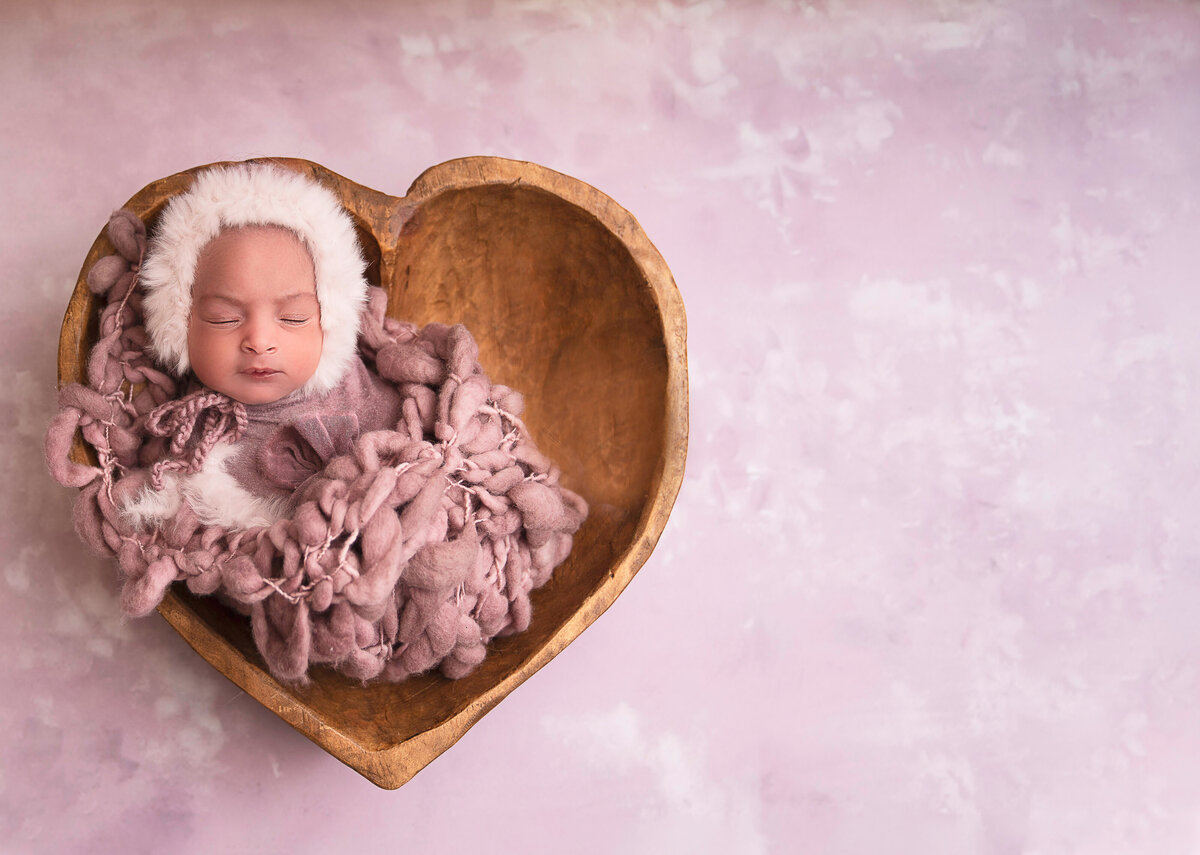 Newborn baby girl is swaddled in mauve blankets and sleeping atop of a heart-shaped bowl. Captured by Bryn Mwar Philadelphia and Main Line photographer Liza Nicole.